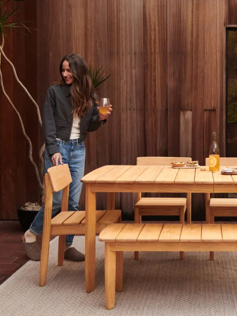 A model standing by a wooden dining set from Burrow, holding a glass of wine. 