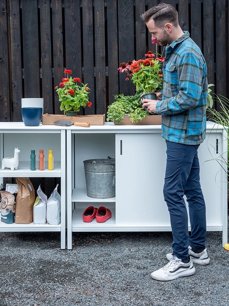 An outdoor storage cupboard with a model standing in front of it, holding a potted plant, from Loll Designs. 