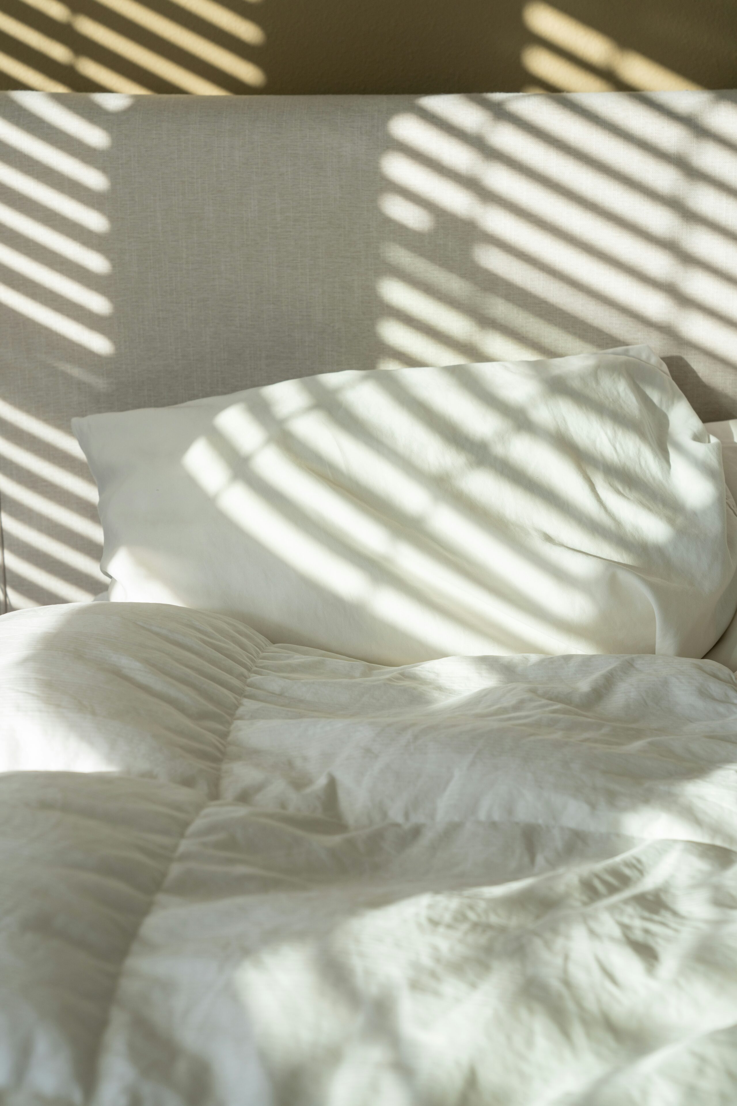 Close up shot of a slept-on bed with white sheets and a white comforters, with the shadows of windows blinds cascading over the bed.
