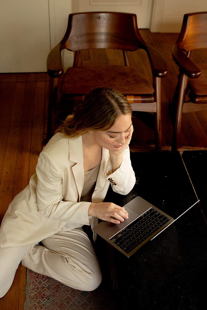 A person in a light-colored suit sits on the floor, working on a laptop in a room with wooden chairs and flooring.
