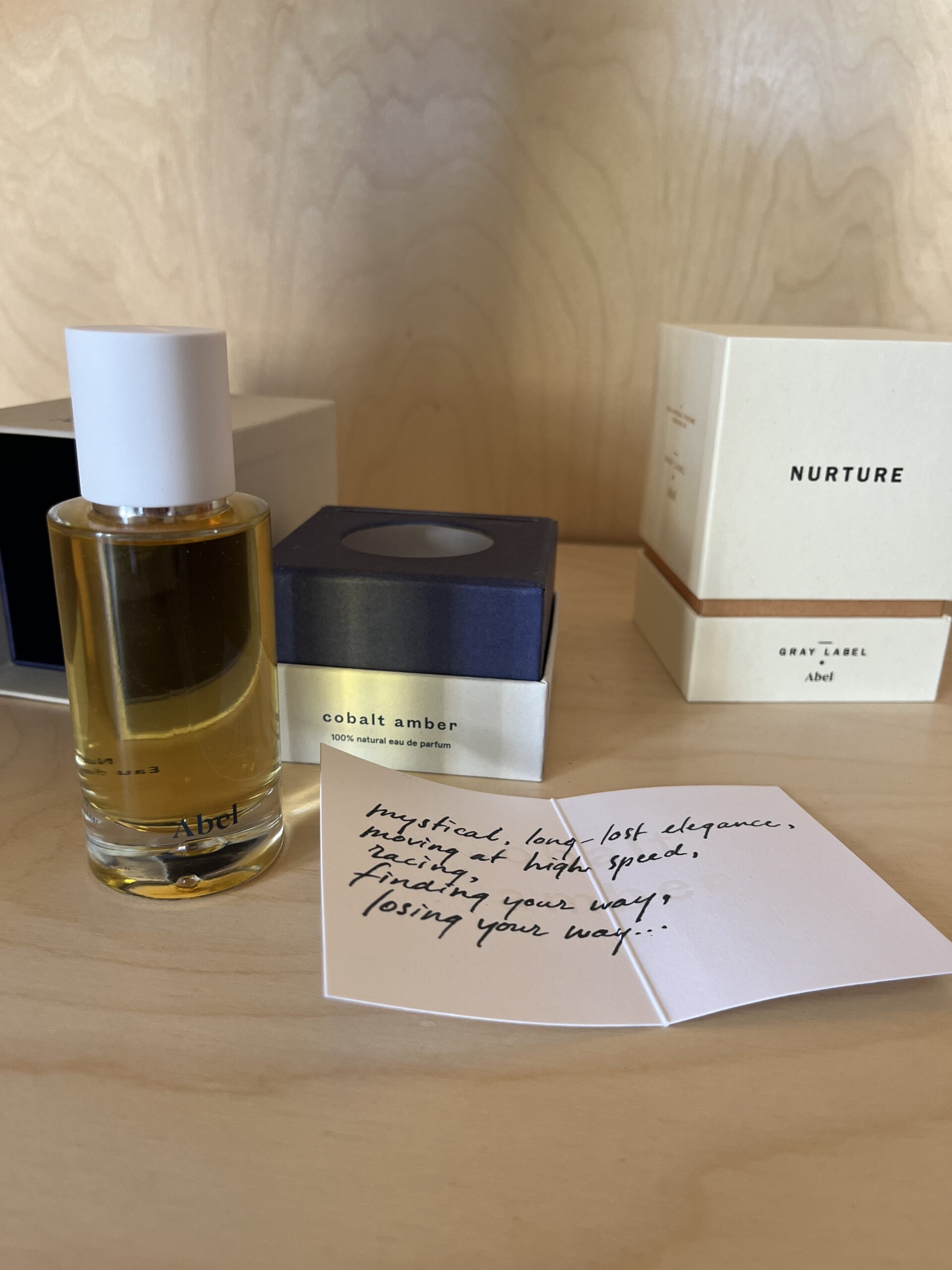 Perfume bottle "Cobalt Amber" with its packaging is next to an open card with handwritten text on a wooden surface. Another box labeled "Nurture" is in the background.