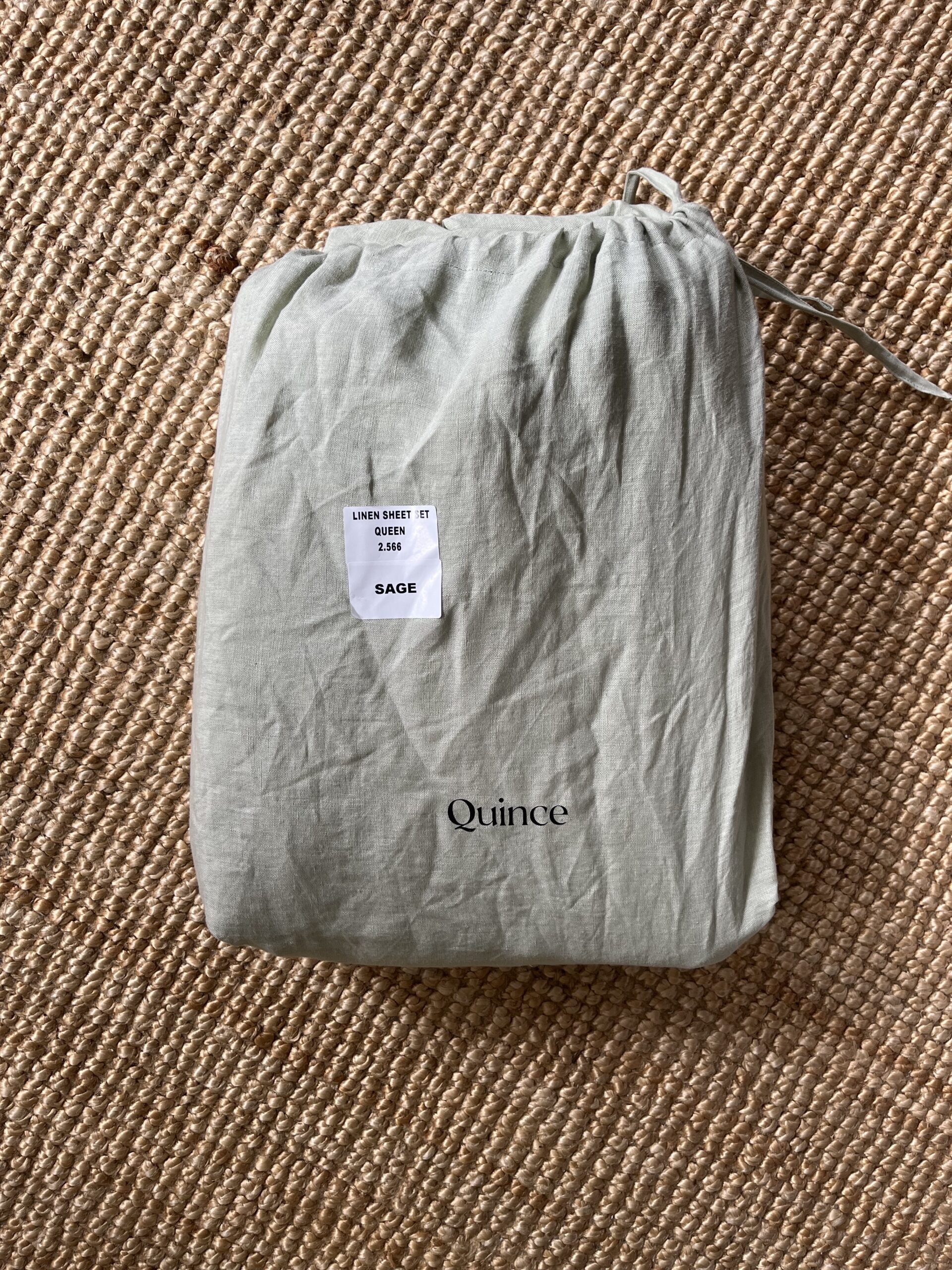 A beige fabric bag with a label reading "Linen Sheet Set - Queen - SAGA" printed in black and another label reading "Quince" is placed on a woven surface.