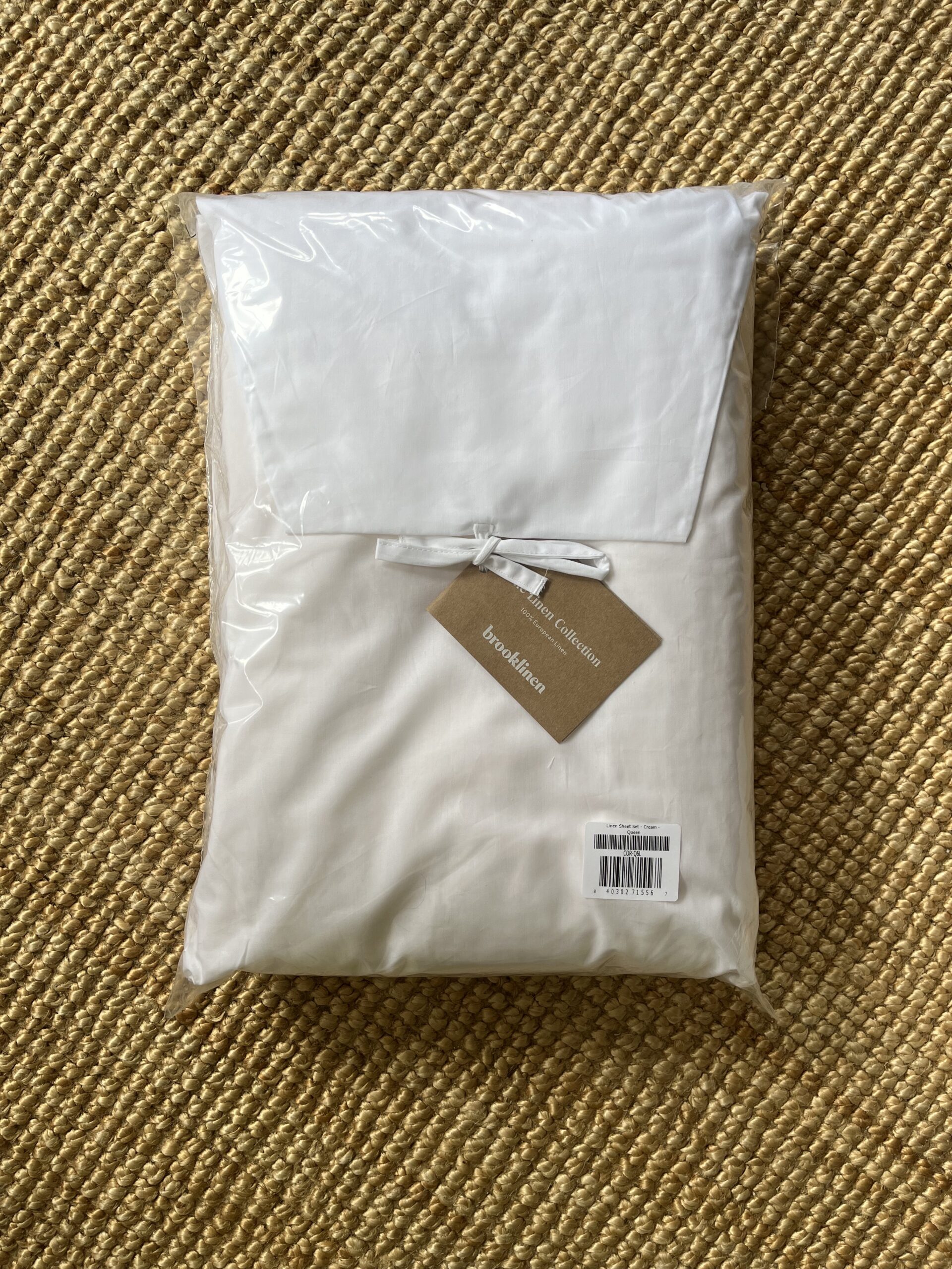 A packaged white and beige bedding set is shown with a cardboard label attached. The set is placed on a woven textured surface.