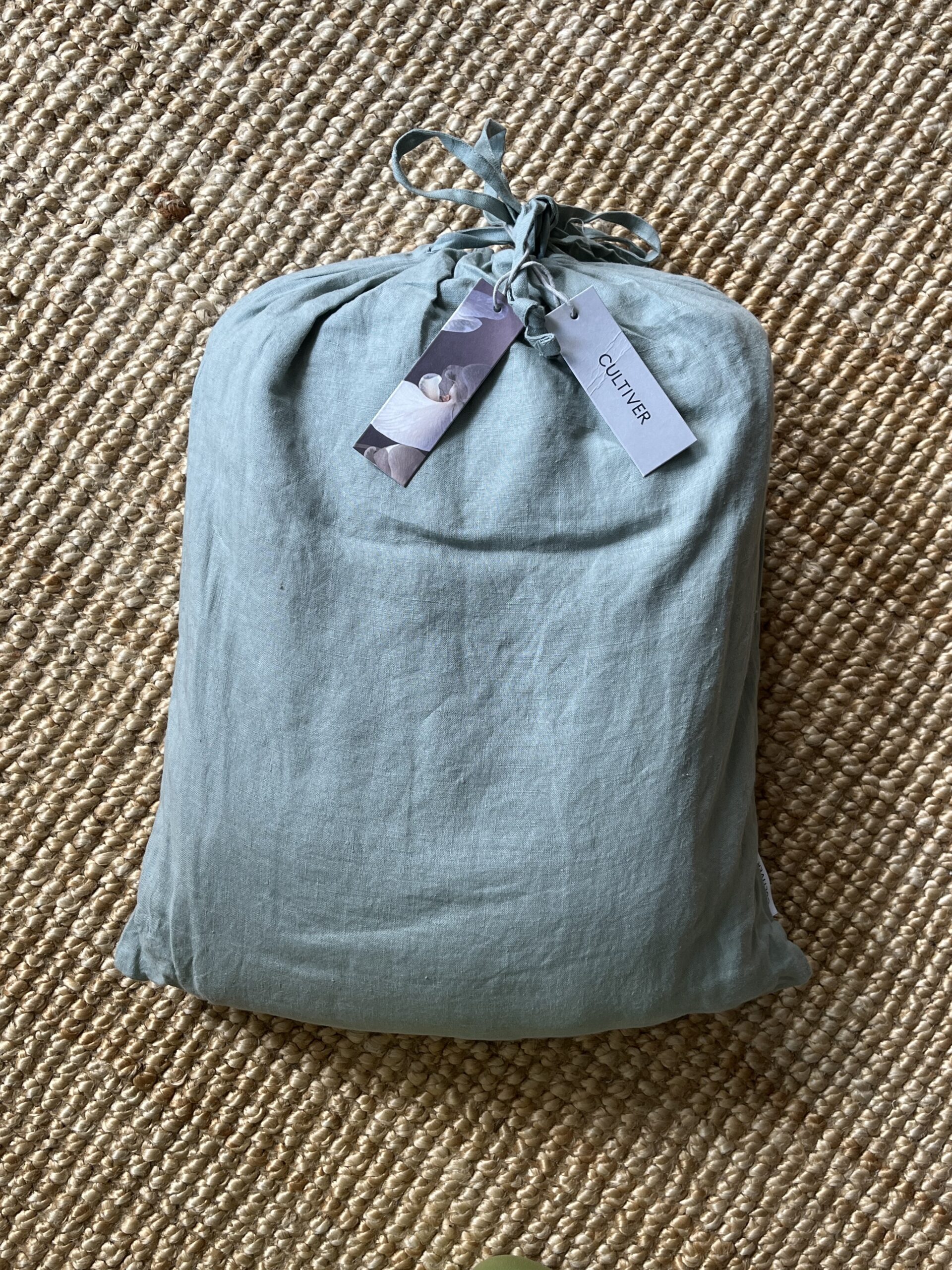 A light green fabric bag tied with a string, labeled "Cultiver," placed on a woven jute rug.