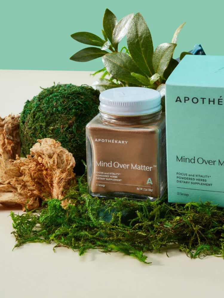 A jar and a box of Apothékary’s “Mind Over Matter” supplement are displayed on a surface with dried mushrooms, moss, and greenery in the background.