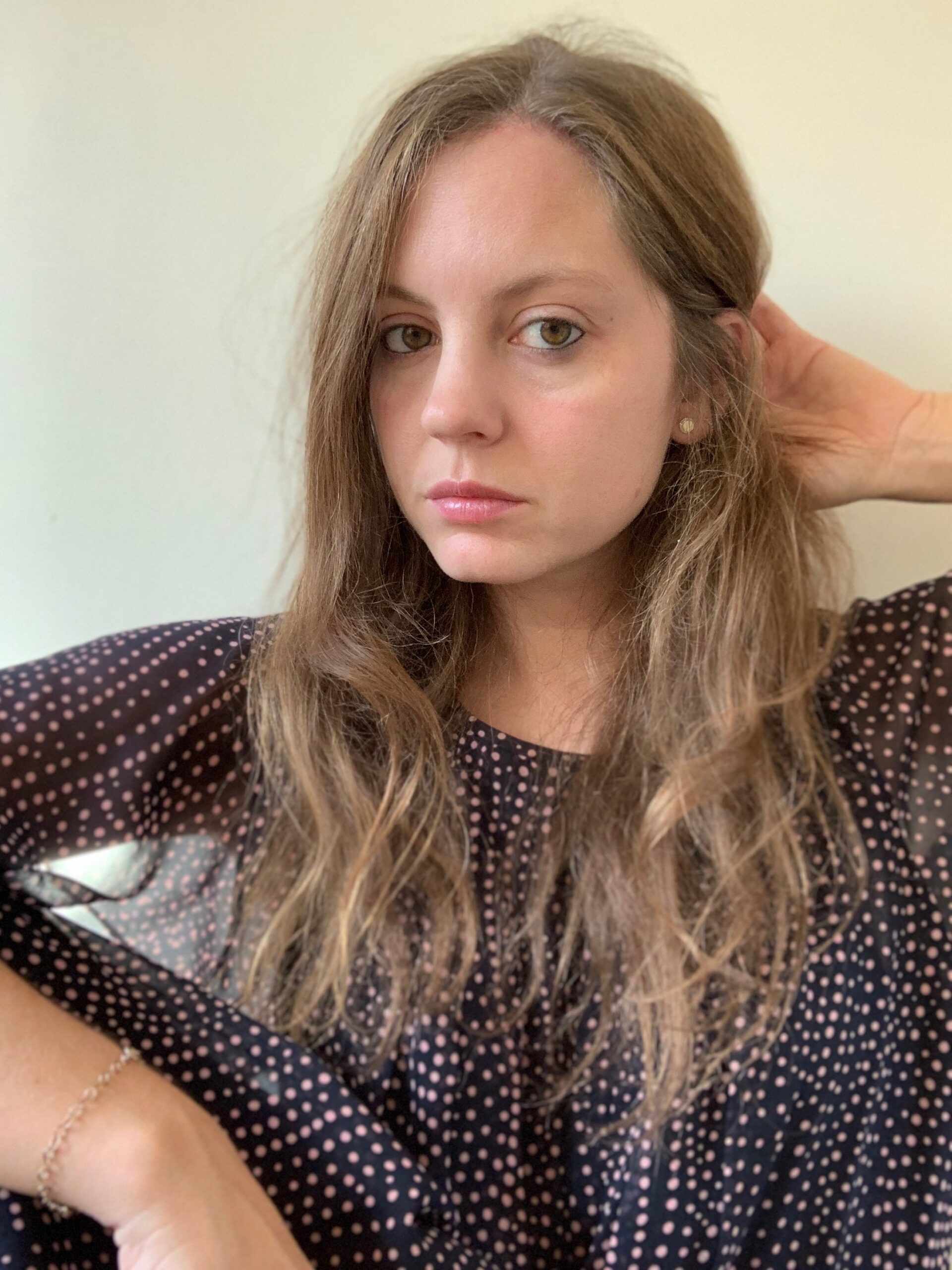 A woman with long, wavy brown hair wearing a black polka dot shirt looks at the camera while touching her hair. She has a neutral expression and is positioned against a plain background.