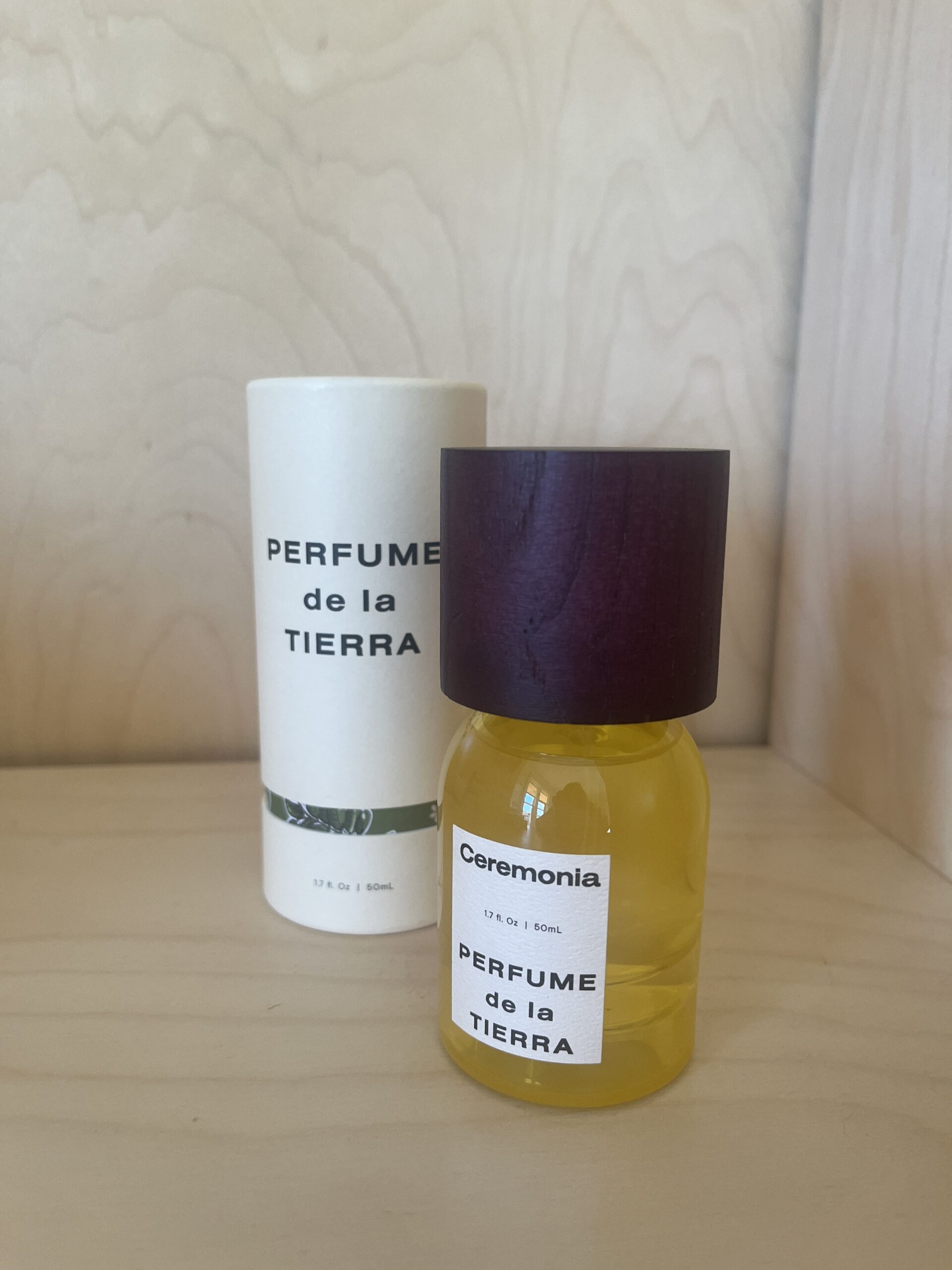 A bottle of "Perfume de la Tierra" by Ceremonia with a wooden cap is in the foreground. The packaging box is in the background on a wooden surface.