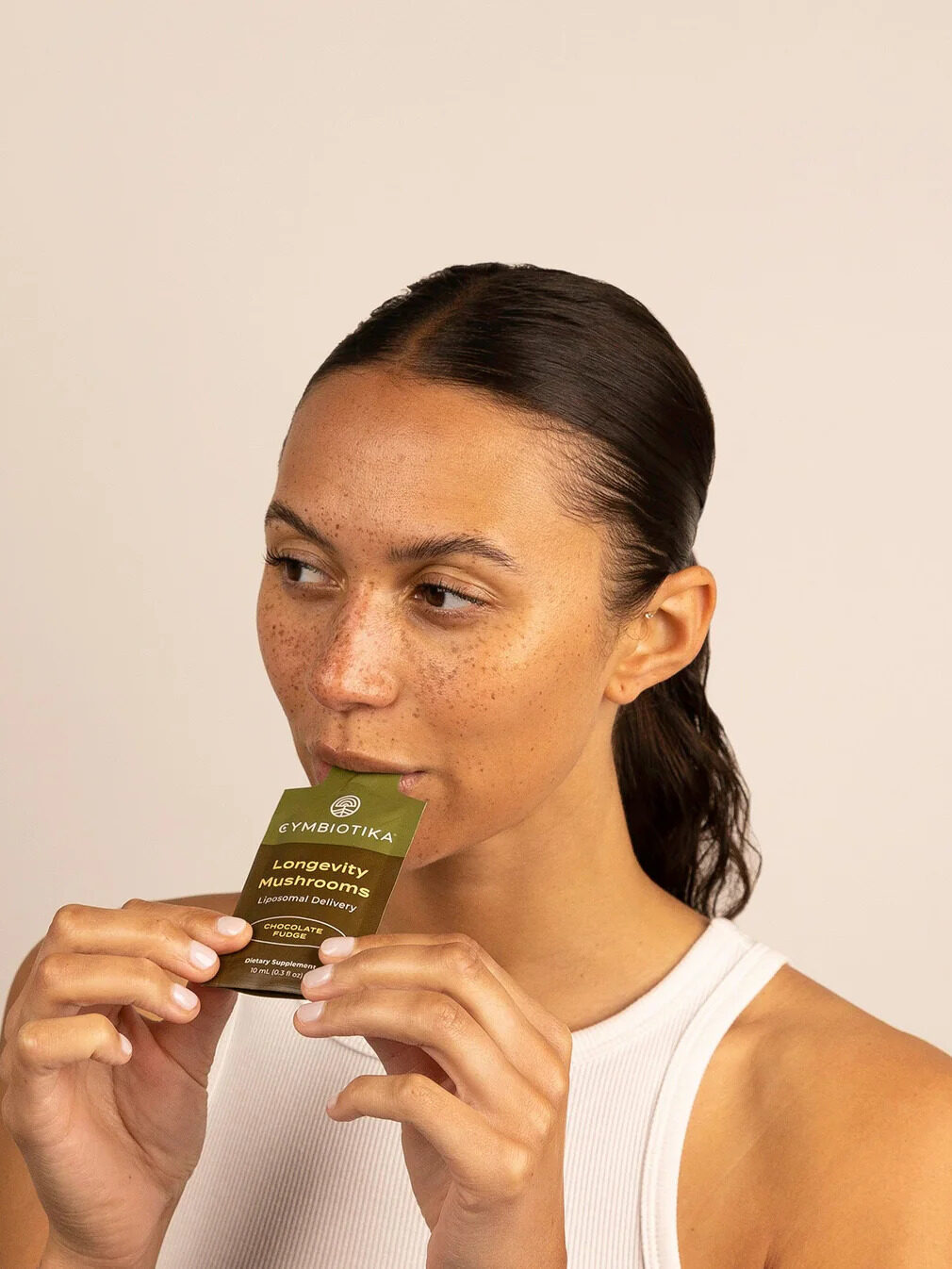 Person in a white tank top holds a "Longevity Mushroom" supplement sachet from Cymbiotika close to their mouth.