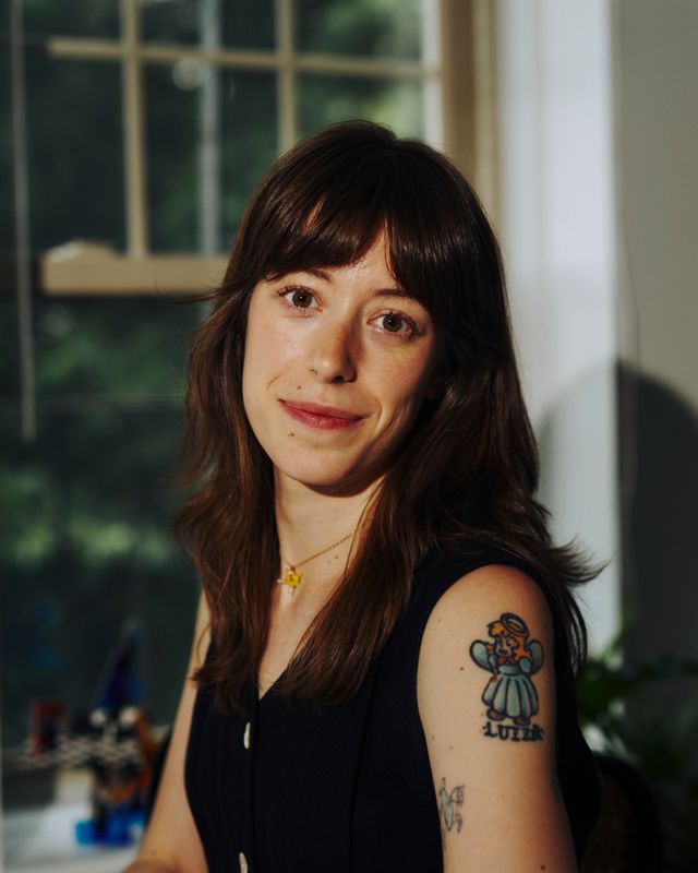 A woman with long brown hair and bangs, wearing a sleeveless dark top, displays a tattoo of a cartoon character with the name "Luiza" on her upper arm. She is smiling, with a window in the background.