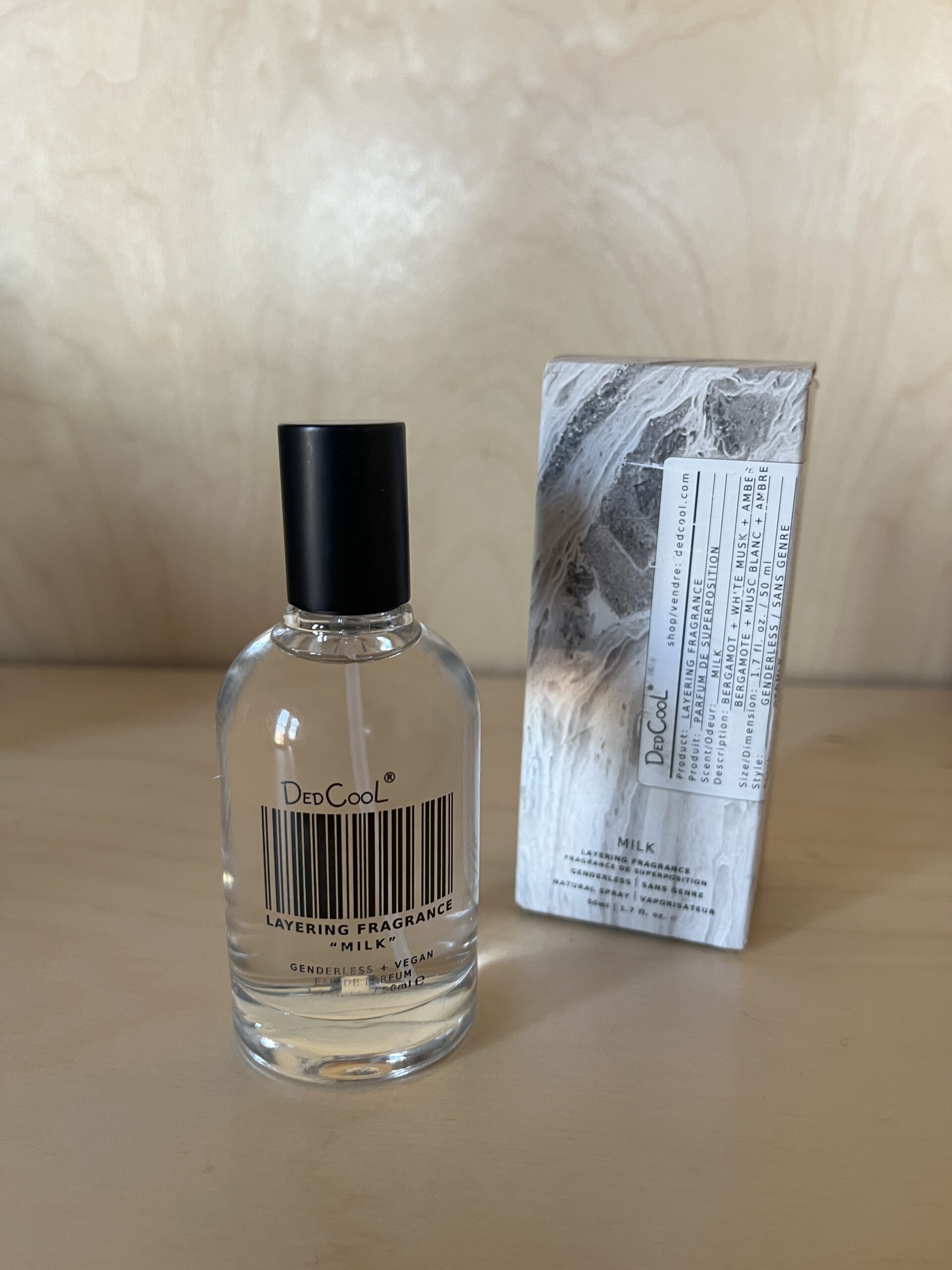 A bottle of DedCool Milk fragrance with a barcode design on the label stands next to its box on a wooden surface.