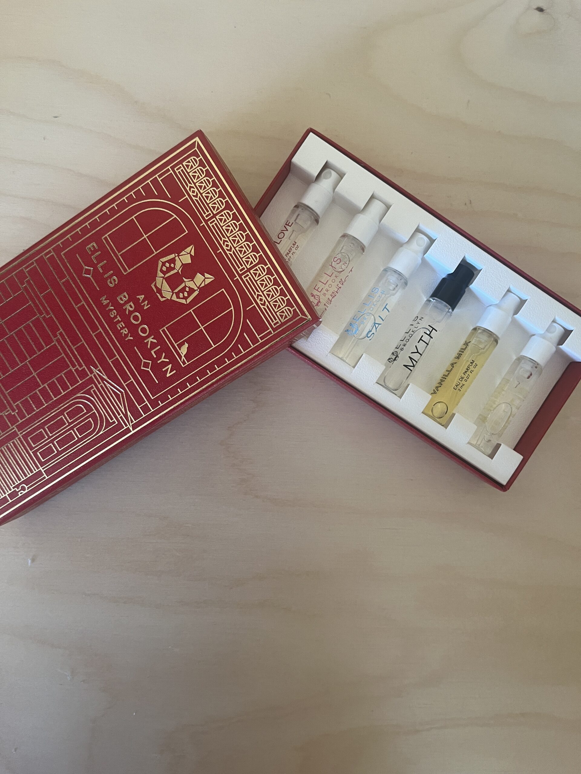 A red box containing six assorted perfume sample vials displayed on a light wood surface.