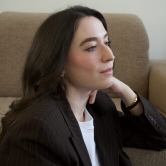 A woman with long dark hair sits on a beige sofa, looking to the side. She wears a dark striped blazer, a white top, hoop earrings, and a bracelet.