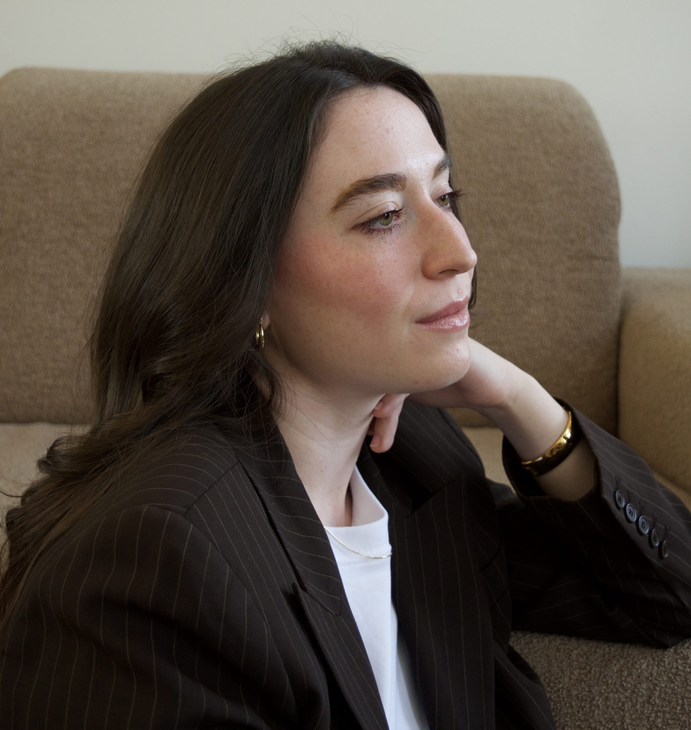 A woman with long dark hair sits on a beige sofa, looking to the side. She wears a dark striped blazer, a white top, hoop earrings, and a bracelet.