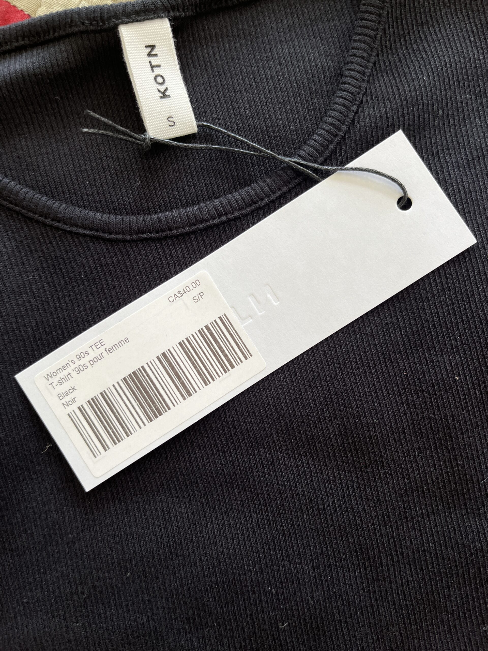 Close-up of a black ribbed T-shirt with a white tag attached, displaying the brand "KOTN" and a barcode. The tag describes the item as "Women's 90s TEE," priced at CA$48.00.