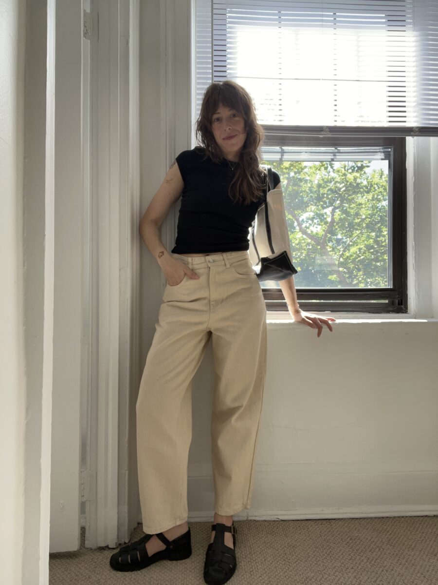 A person wearing a black shirt and beige pants stands inside a room by a window with partially open blinds. They have one arm resting on the windowsill and sunlight is coming through the window.