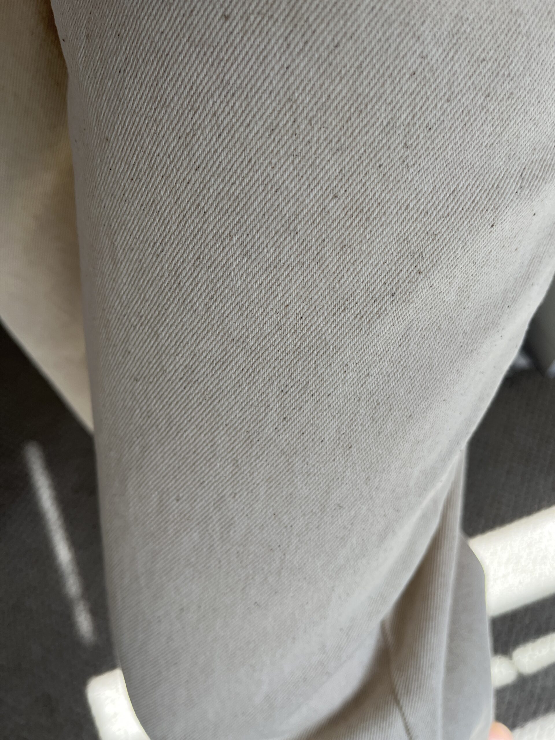 Close-up view of a light-colored, textured fabric with a subtle diagonal pattern. There is a small shadow and portion of a darker surface visible at the bottom right corner.