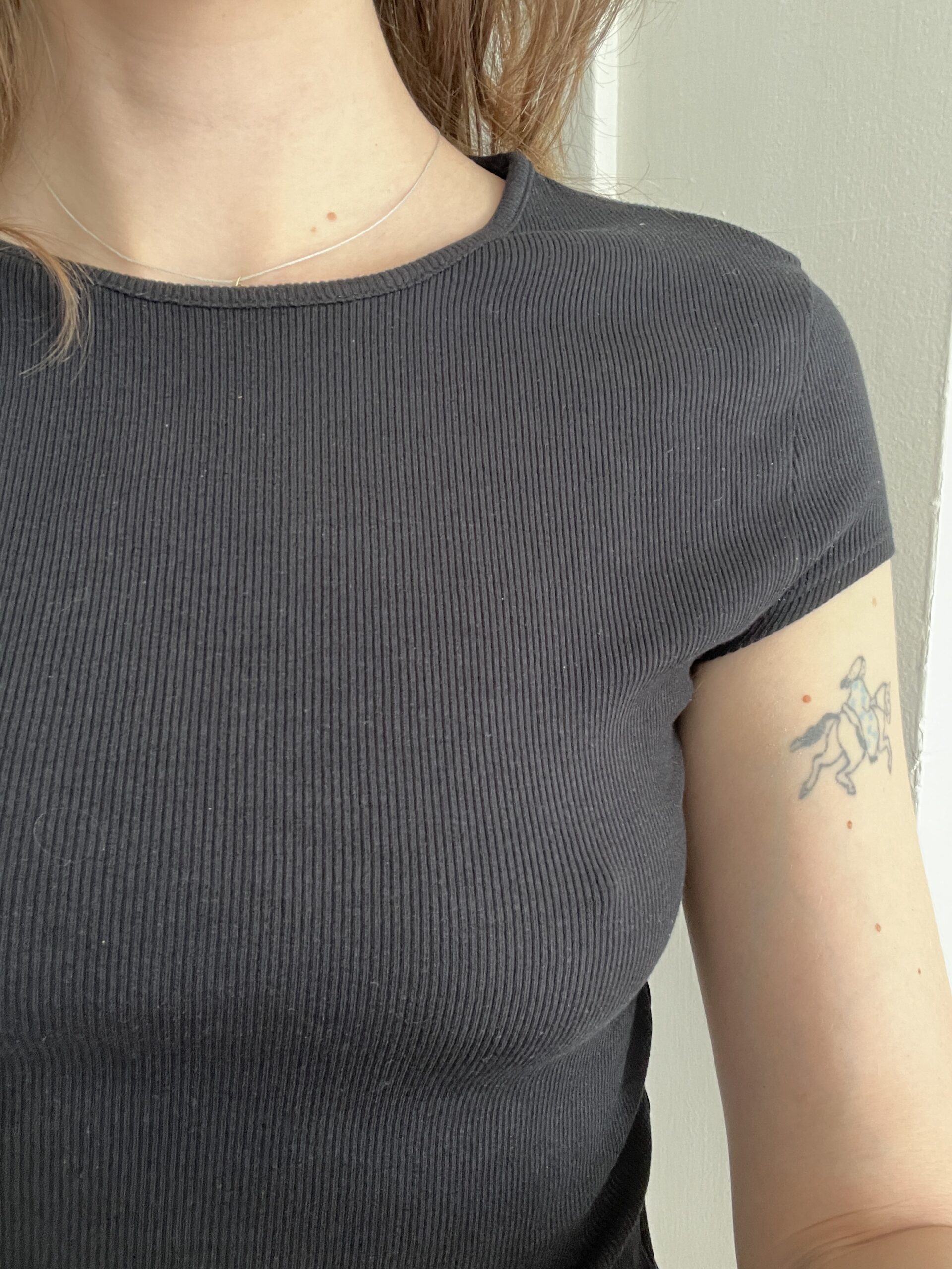 A person wearing a black ribbed short-sleeve top is shown from the neck to the torso, with a tattoo of a small outlined figure on their upper left arm.