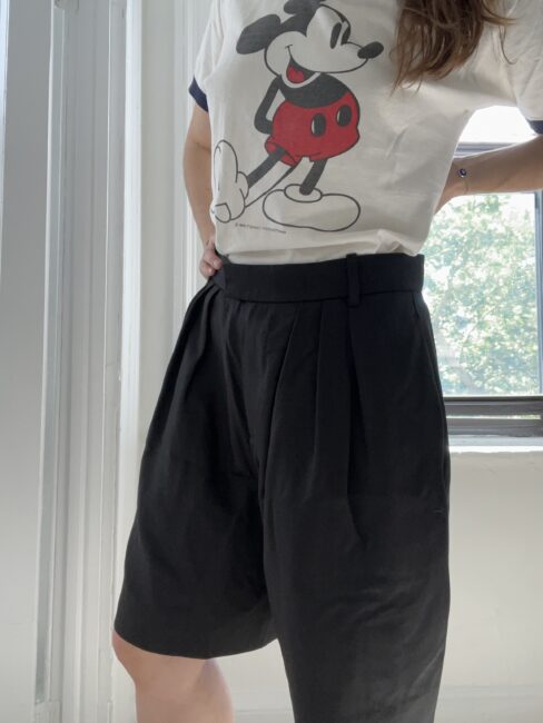 A person is wearing a T-shirt with a classic cartoon character and high-waisted black shorts, standing near a window.