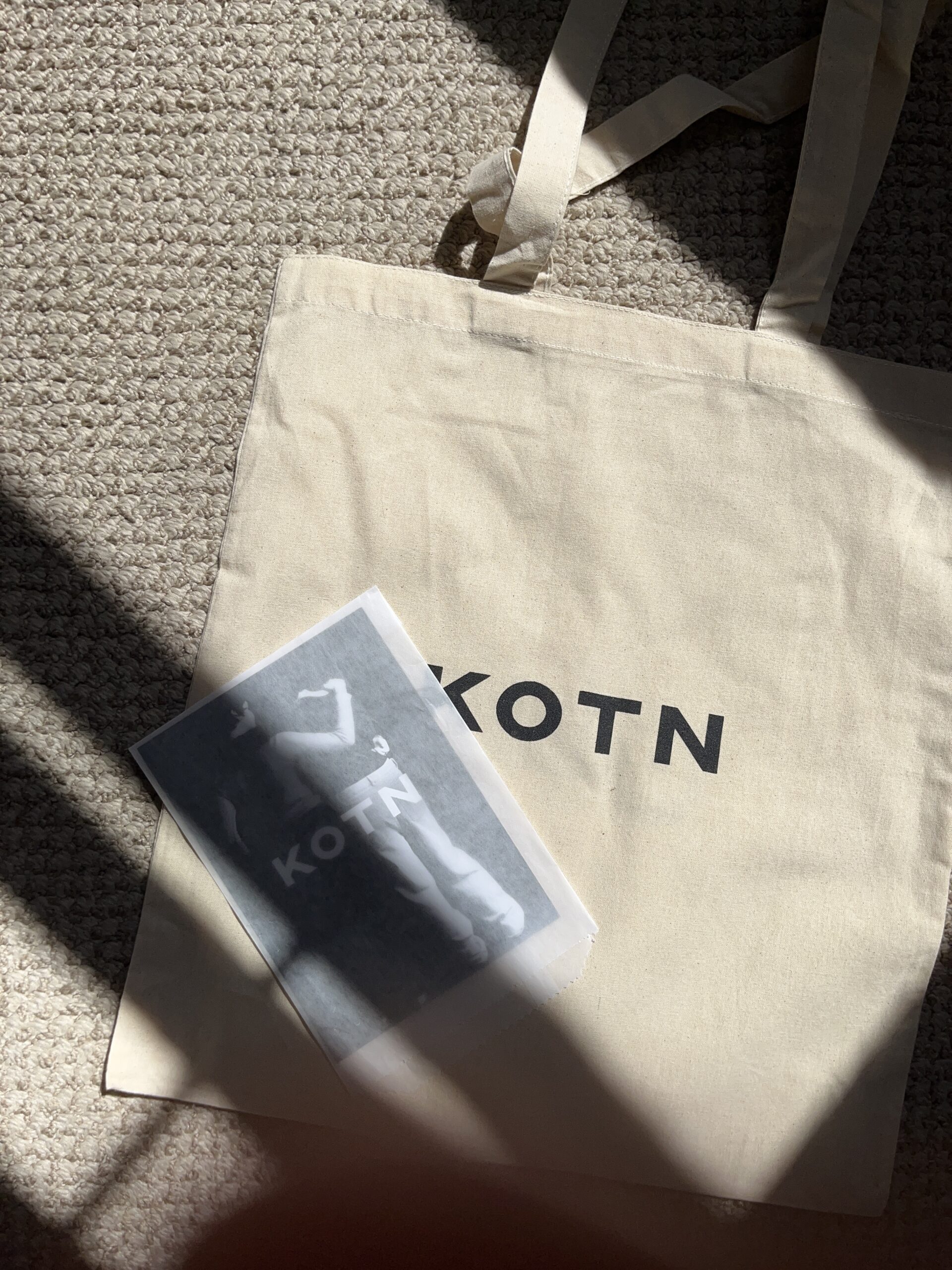 A beige tote bag with "KOTN" printed in black letters lies on a textured floor next to a black and white image partially covered in shadow.