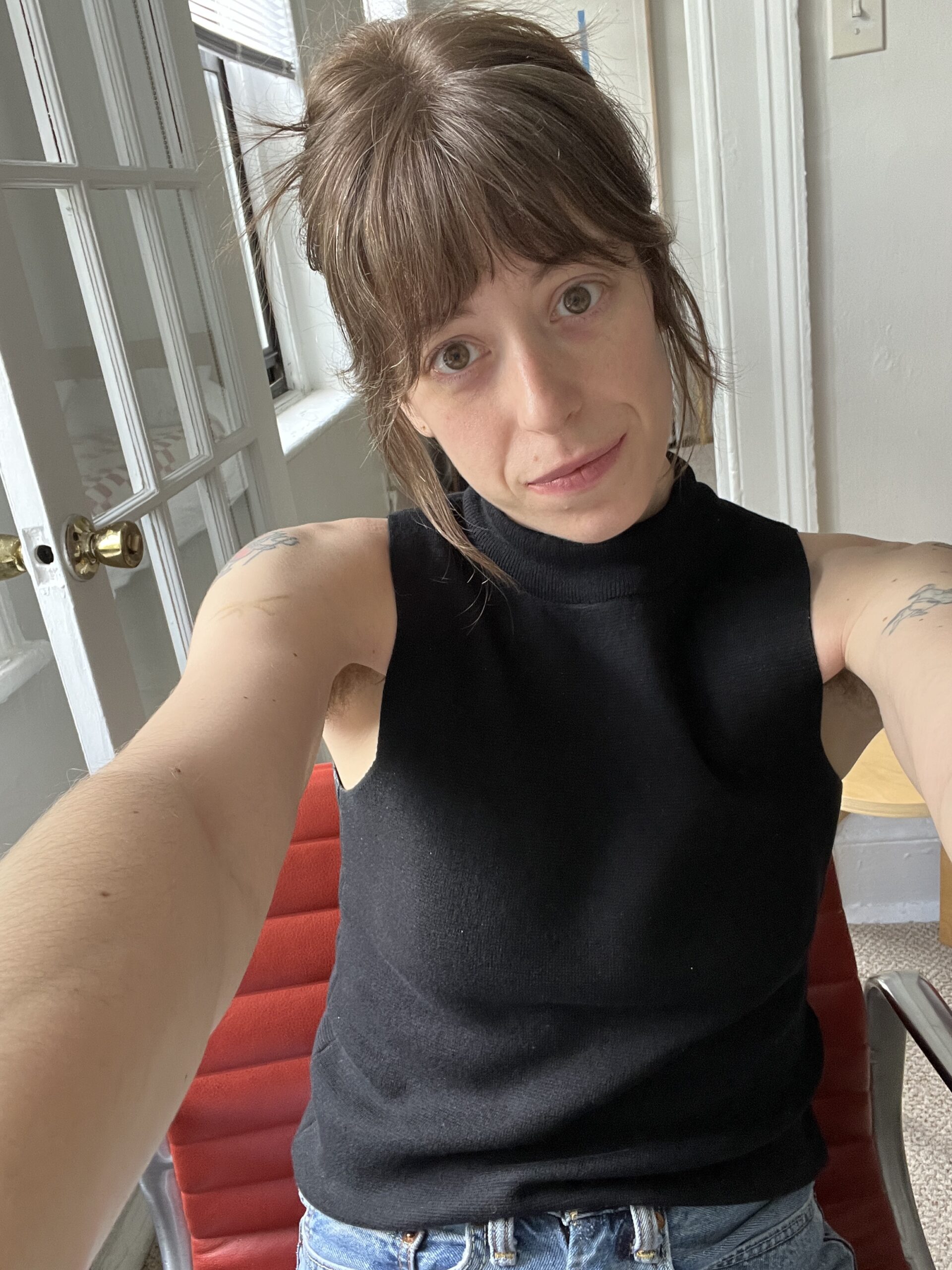 A person with long brown hair wearing a sleeveless black top takes a selfie while sitting on a red chair in a room with white walls and a door with glass panes in the background.