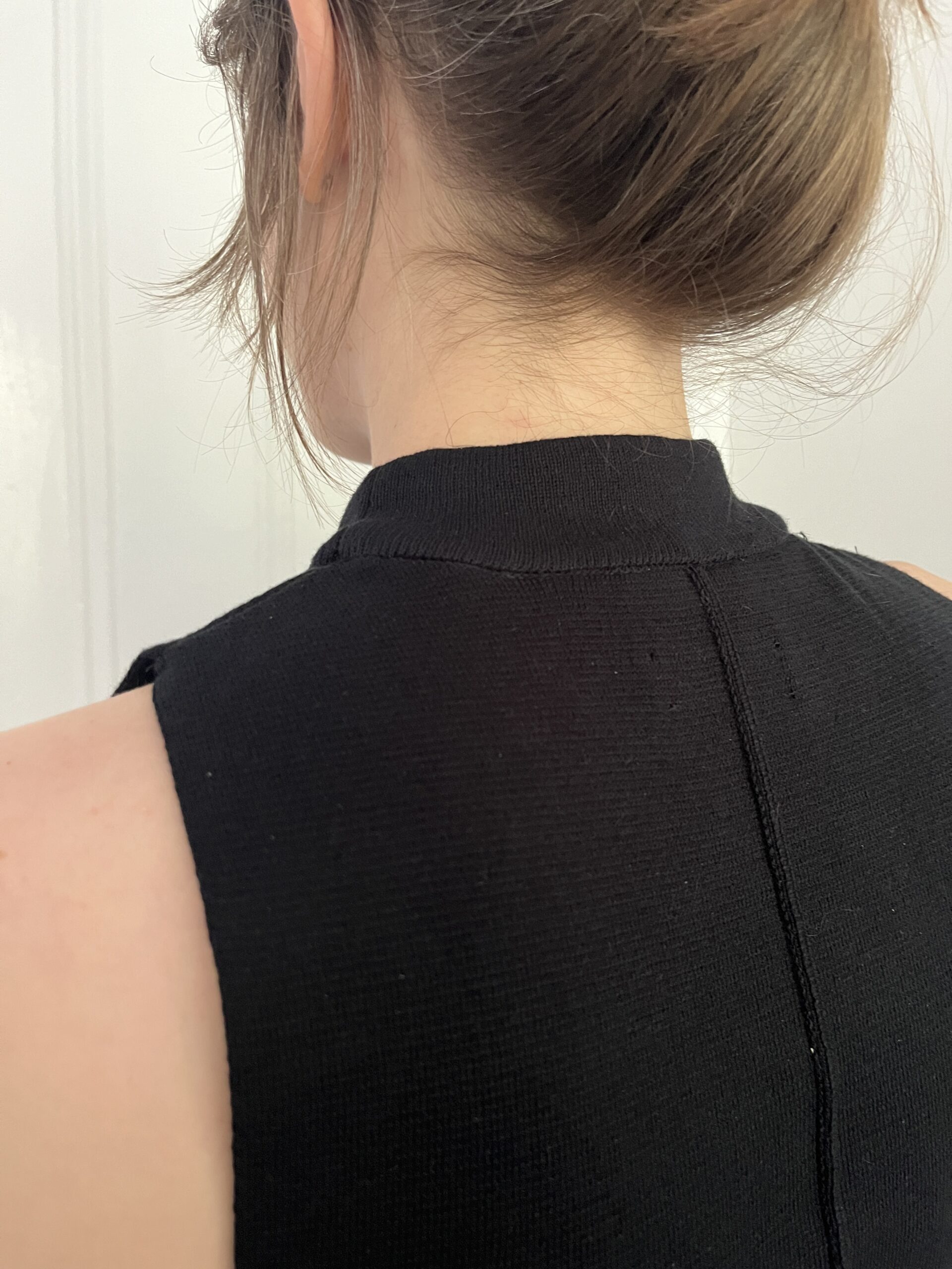 Close-up of the back of a person's head and neck, showing a brown hair bun and a black sleeveless top with a seam running down the back.