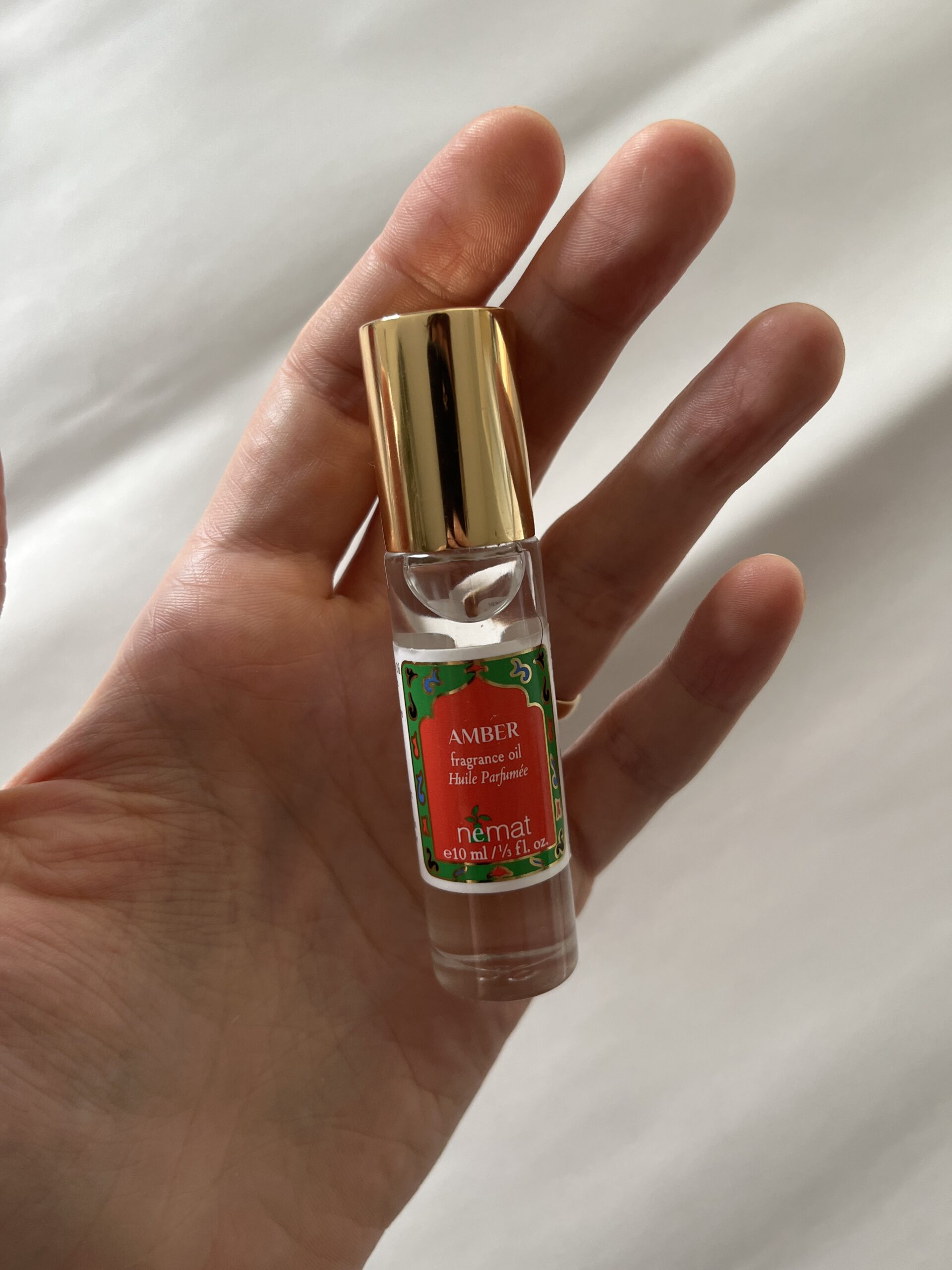 A hand holds a small glass bottle of Amber fragrance oil with a gold cap, labeled "Amber" by Nemat.