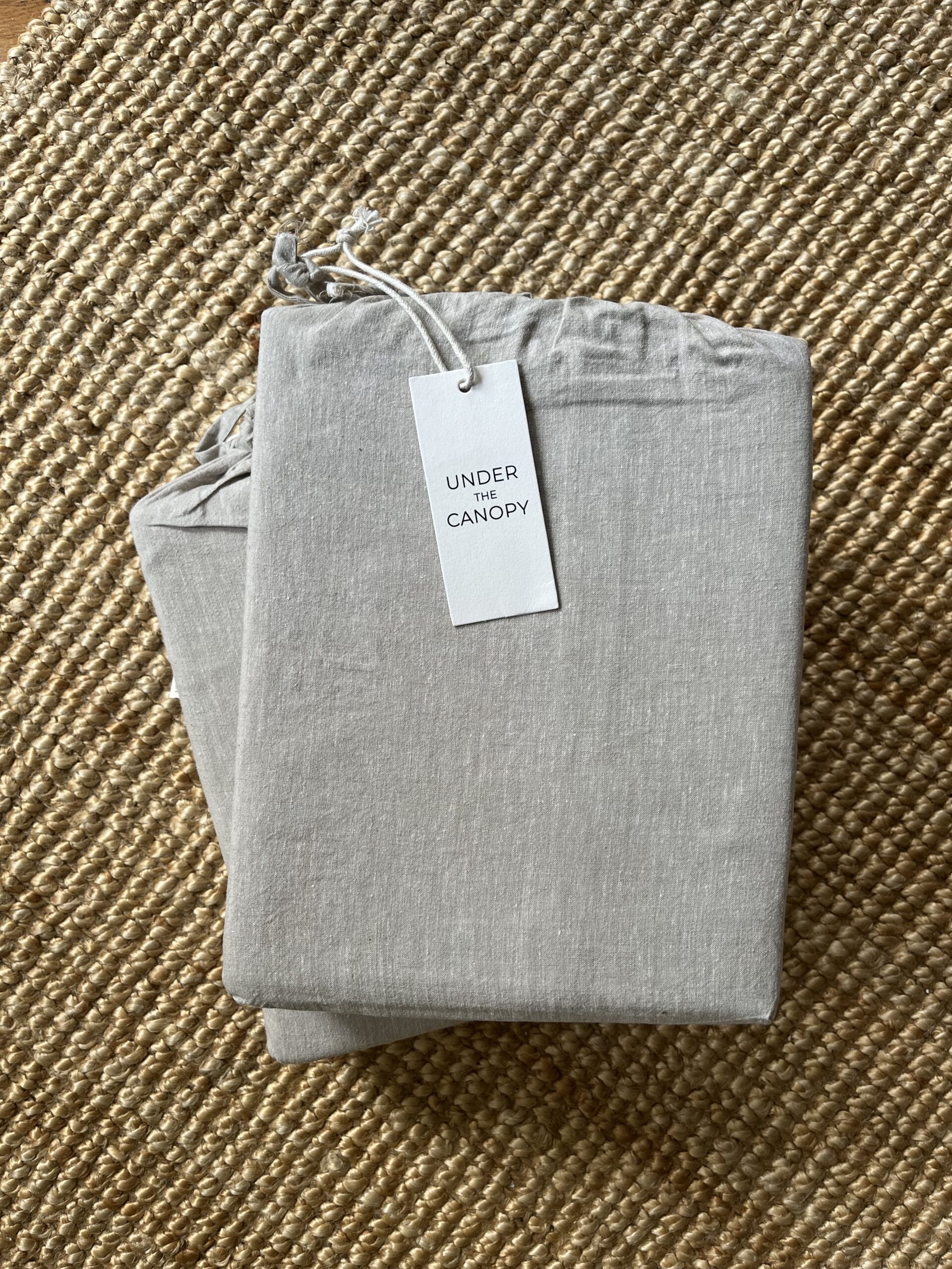 A folded set of grey bed sheets with a tag labeled "Under the Canopy" placed on a woven, beige mat.