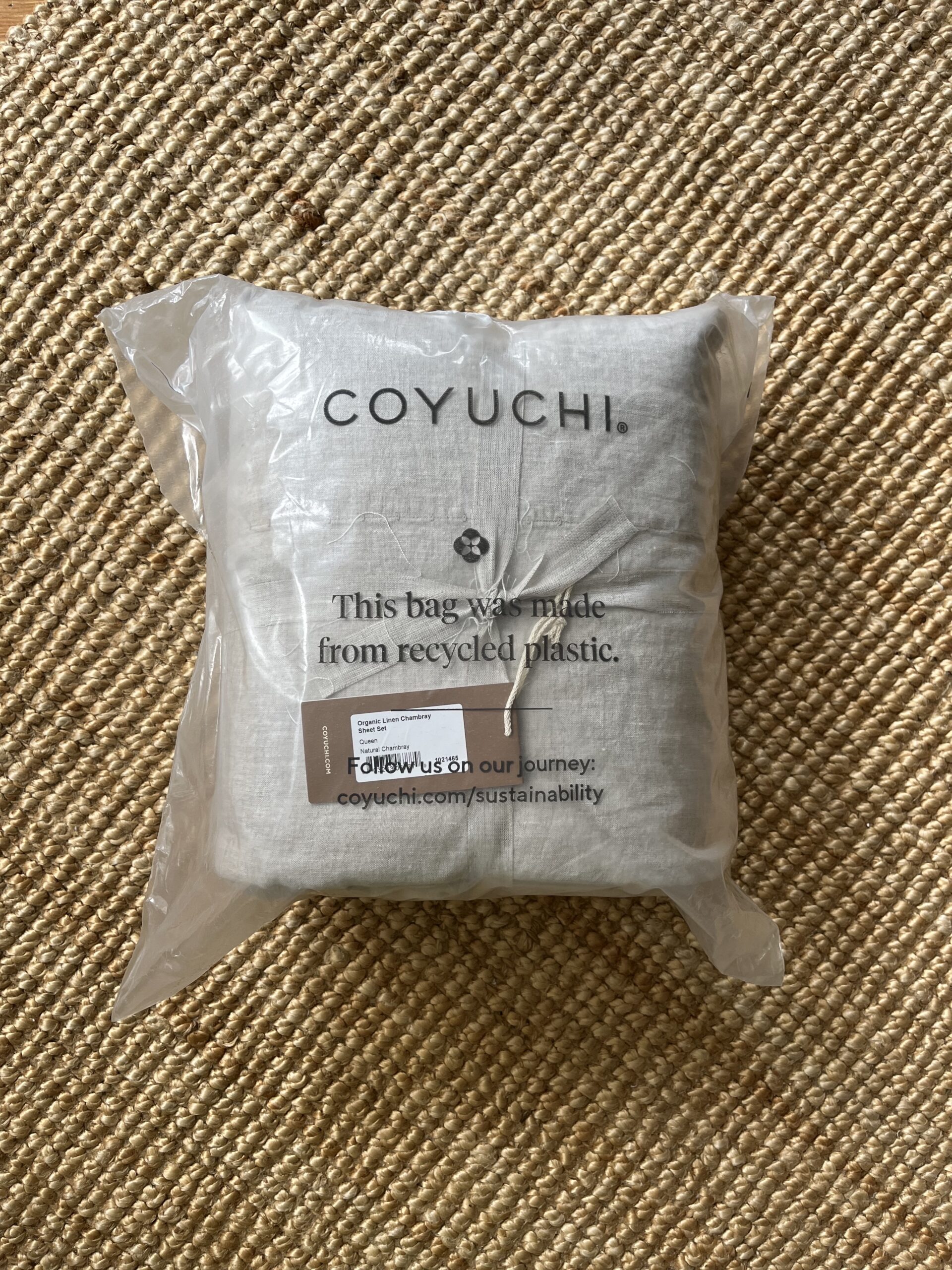 A Coyuchi product in recyclable packaging is placed on a textured mat. The packaging informs that the bag is made from recycled plastic and encourages visiting their website for more sustainability information.