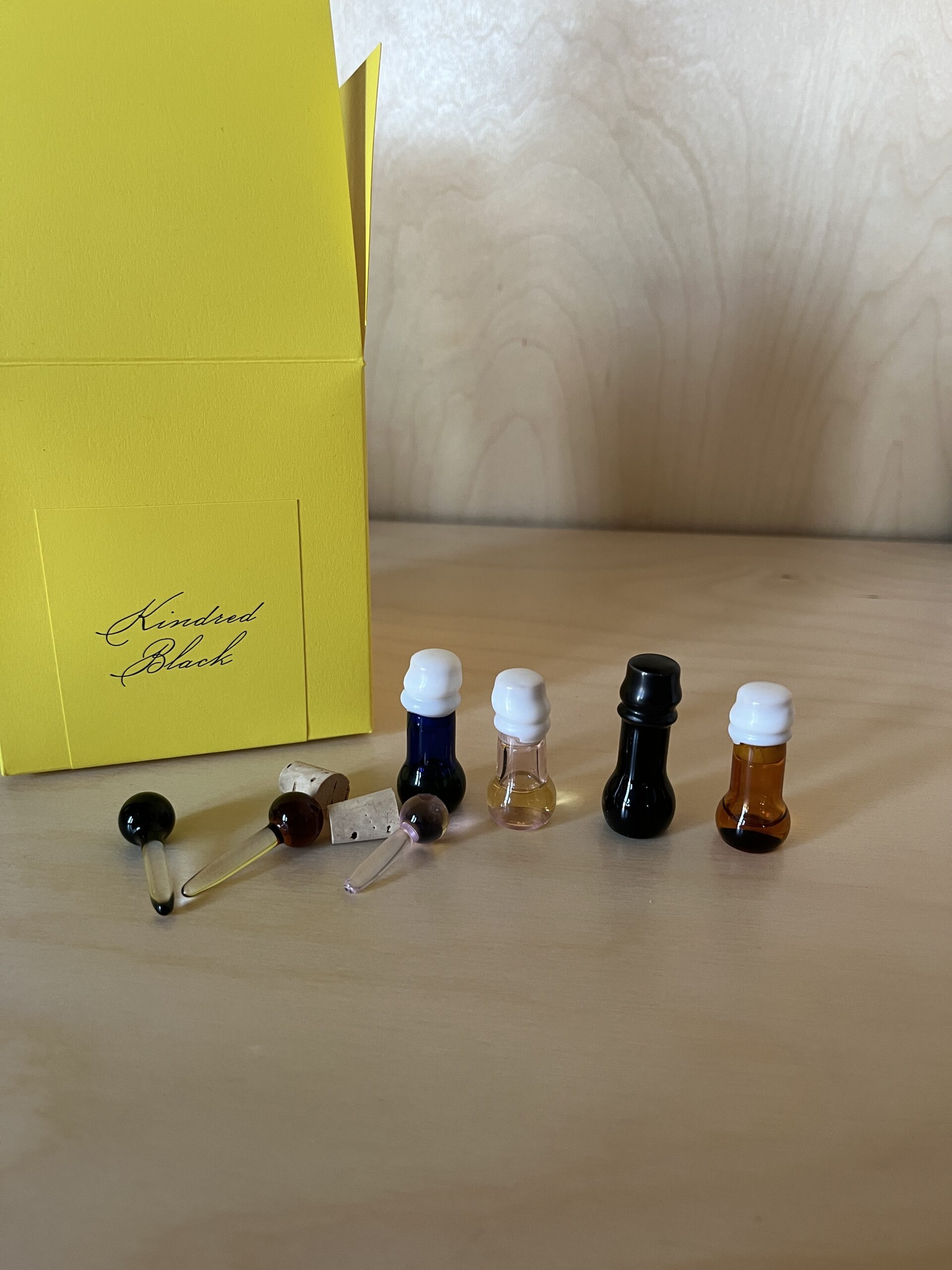 Five small glass bottles with various colored liquids are displayed on a wooden surface, alongside a yellow box labeled "Kindred Black.