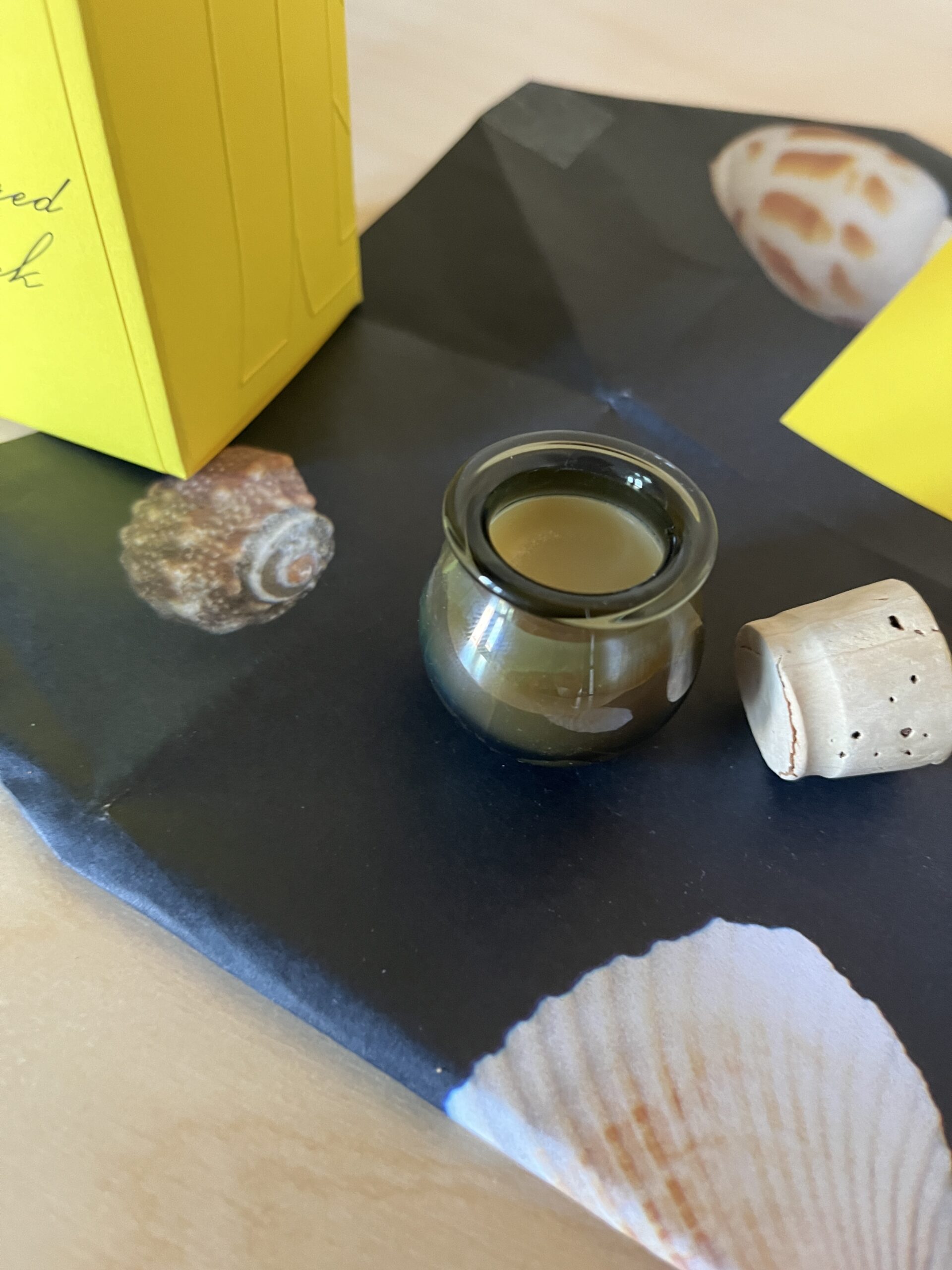 A small glass jar with a cork stopper contains a creamy substance. It is placed on a dark surface decorated with images of seashells. Nearby is a yellow box.