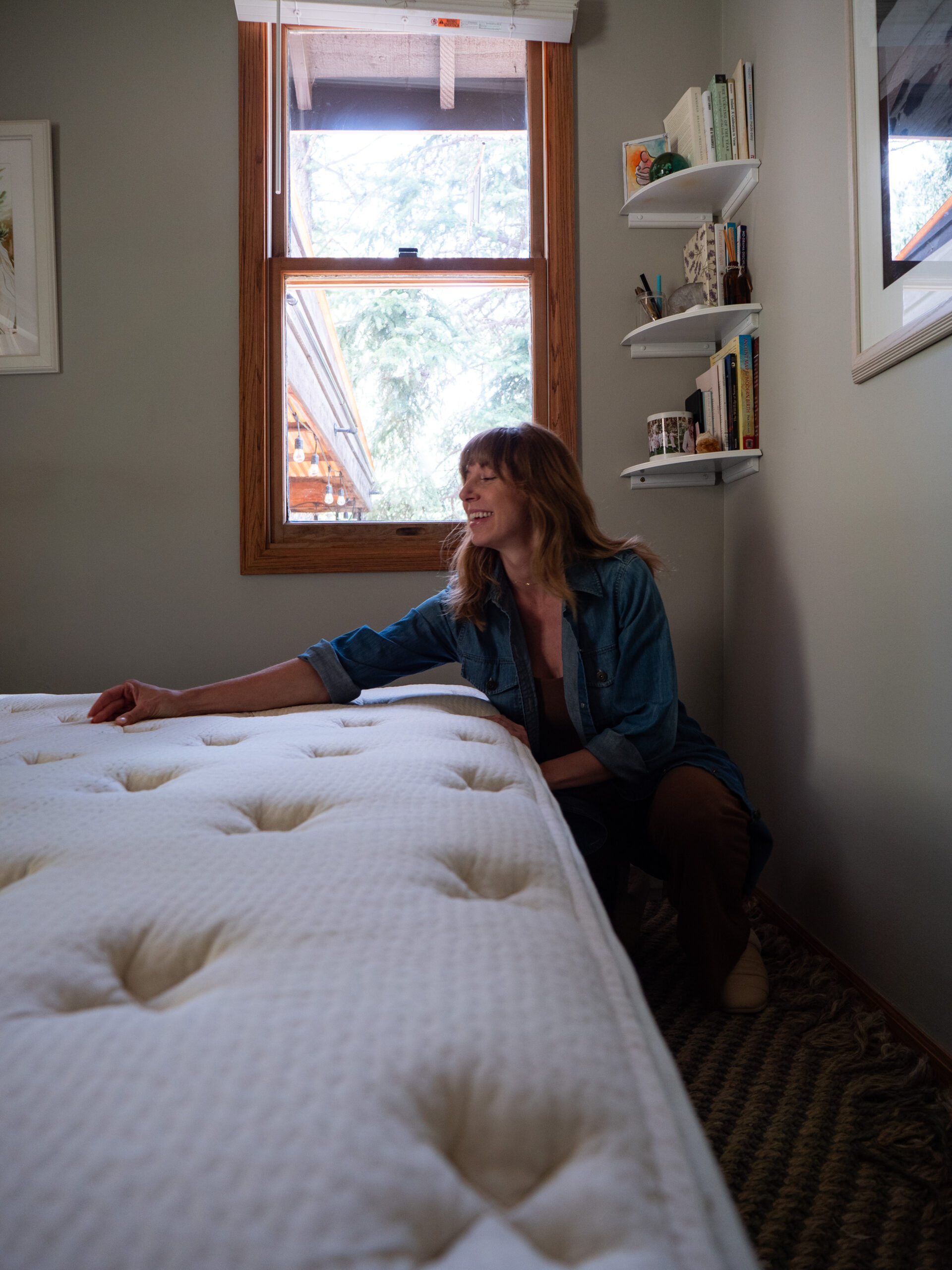 A person smiling and touching the side of a white mattress in a small room with a window, shelves of books, and framed pictures on the wall.