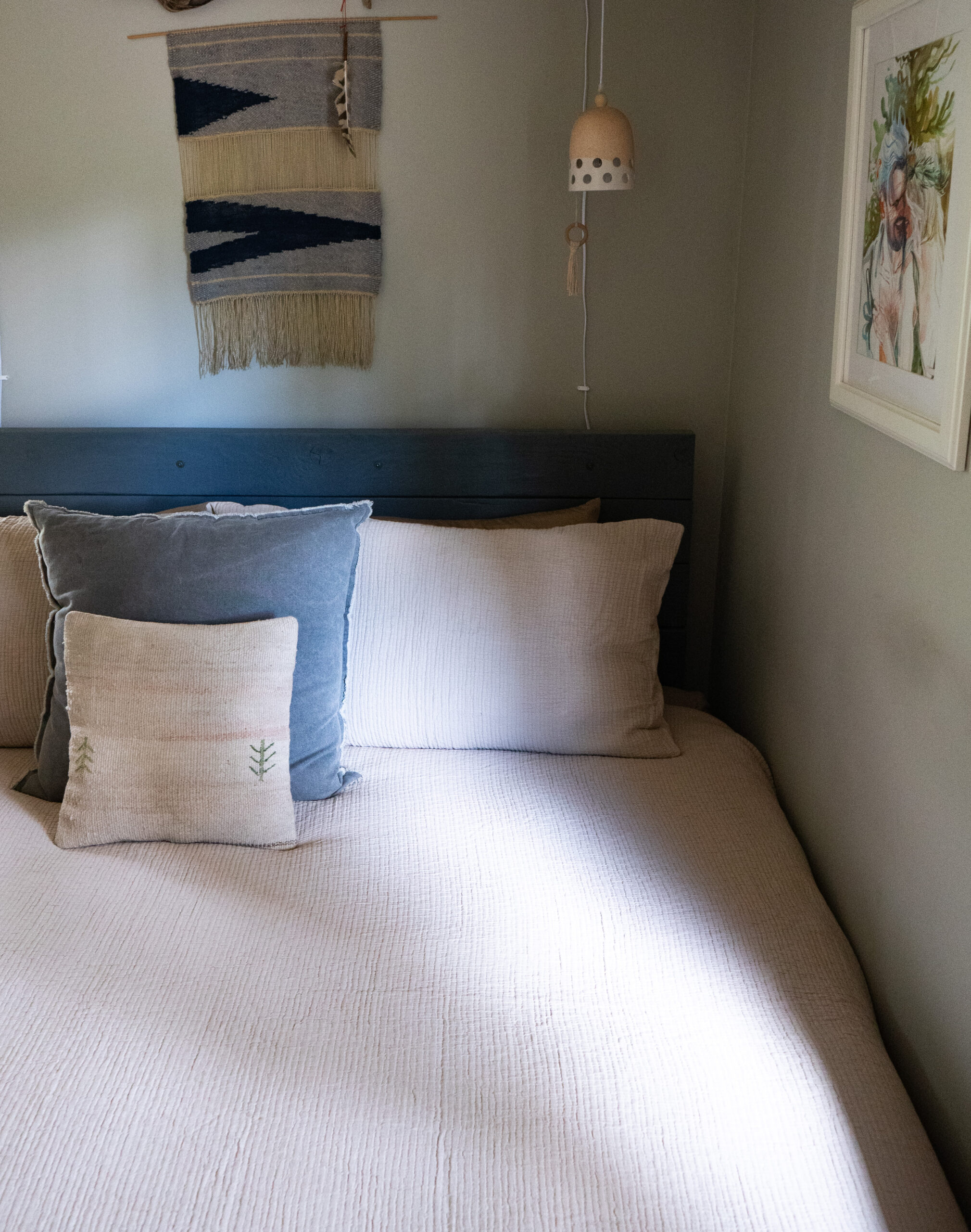 A neatly made bed with white bedding, adorned with two pillows, one white and one gray. On the wall above the bed, there is a woven wall hanging, a pendant light, and a framed picture.