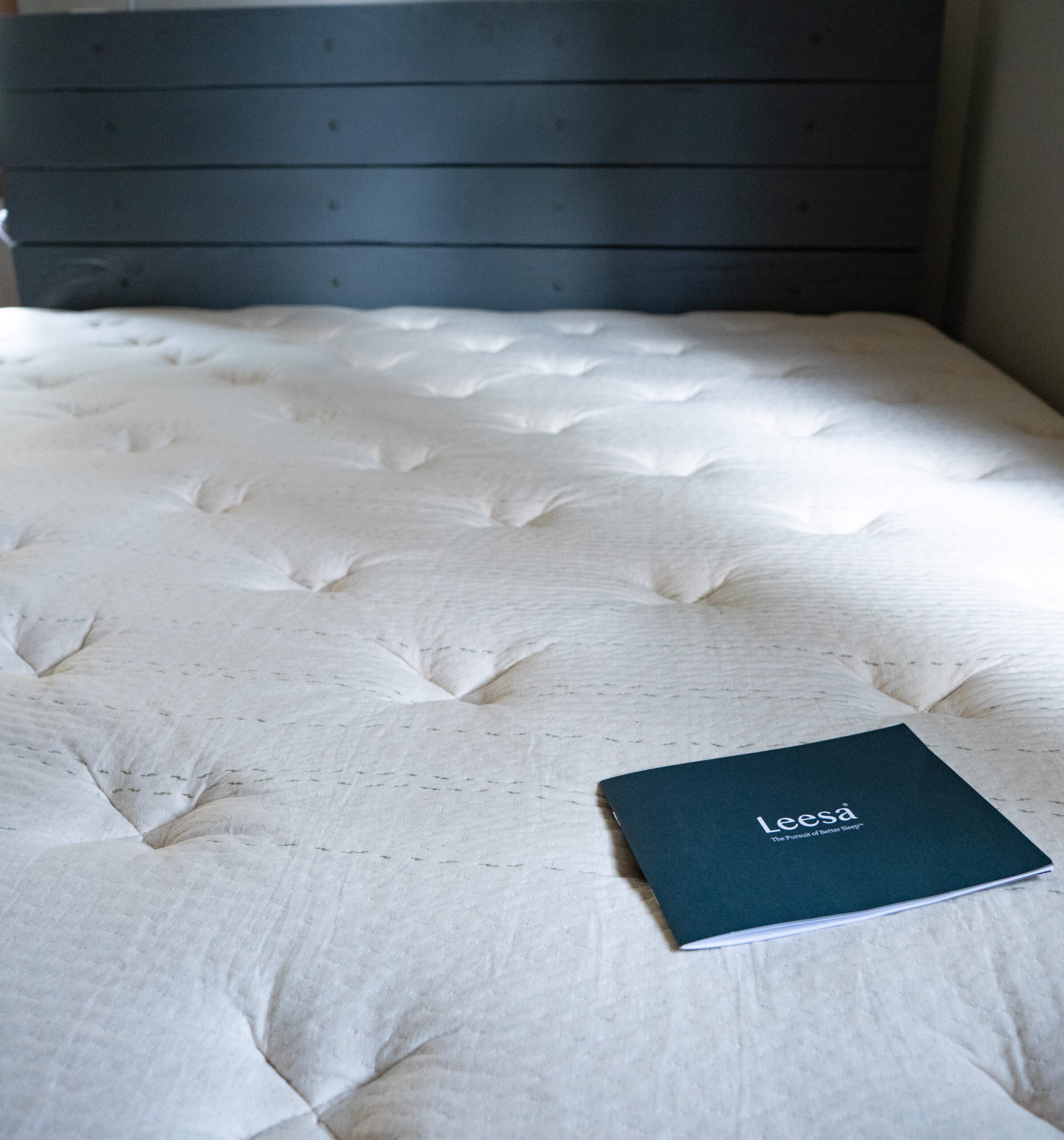 A beige mattress with a booklet labeled "Leesa" placed on top, set against a dark wooden headboard.