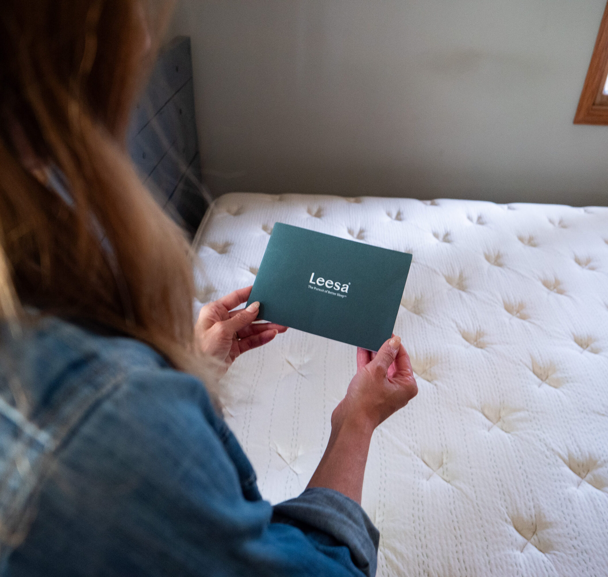 A person holding a Leesa mattress information card while facing a white mattress in a bedroom.