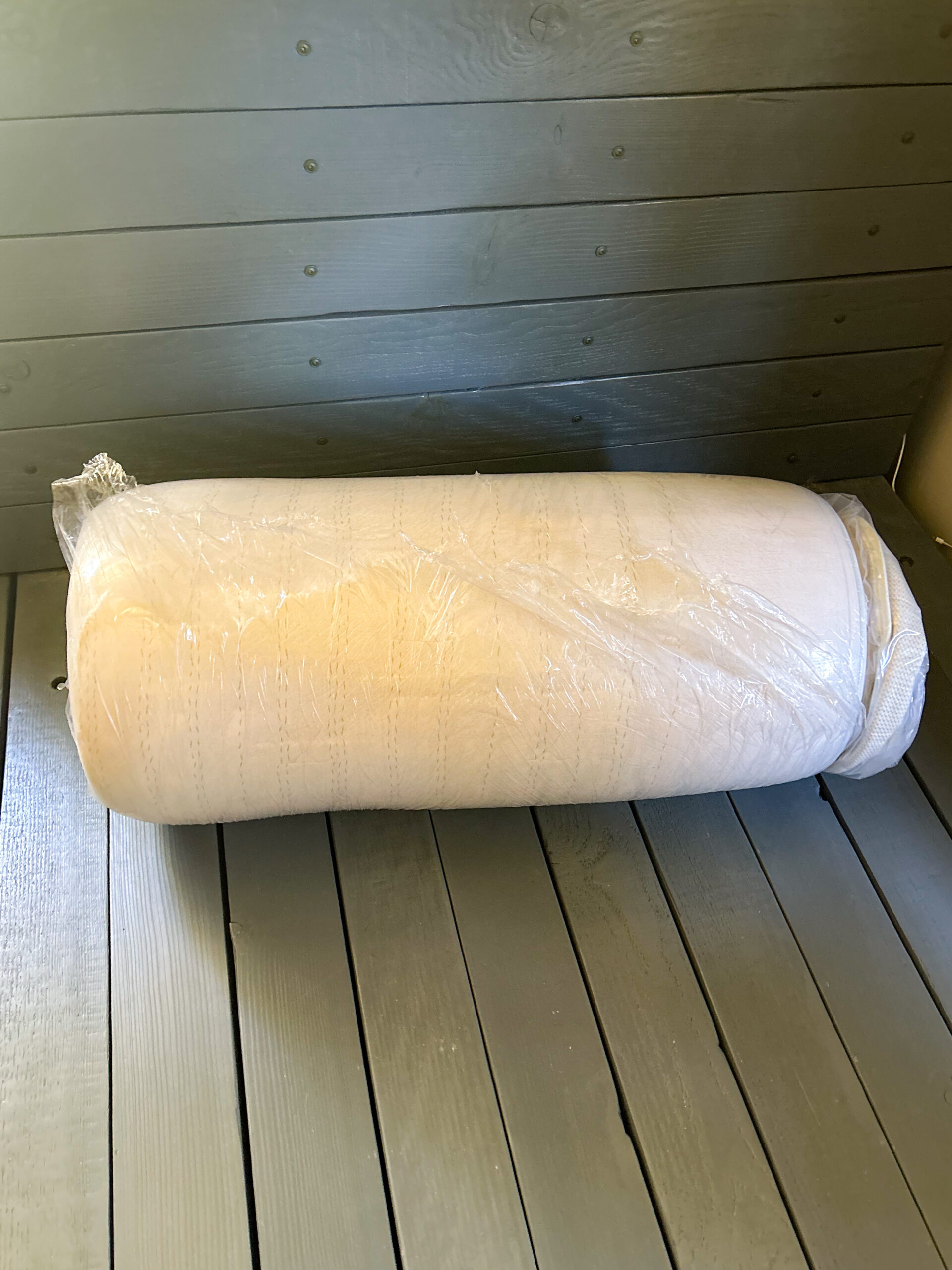 A rolled-up white mattress is wrapped in plastic and placed on a gray wooden floor.