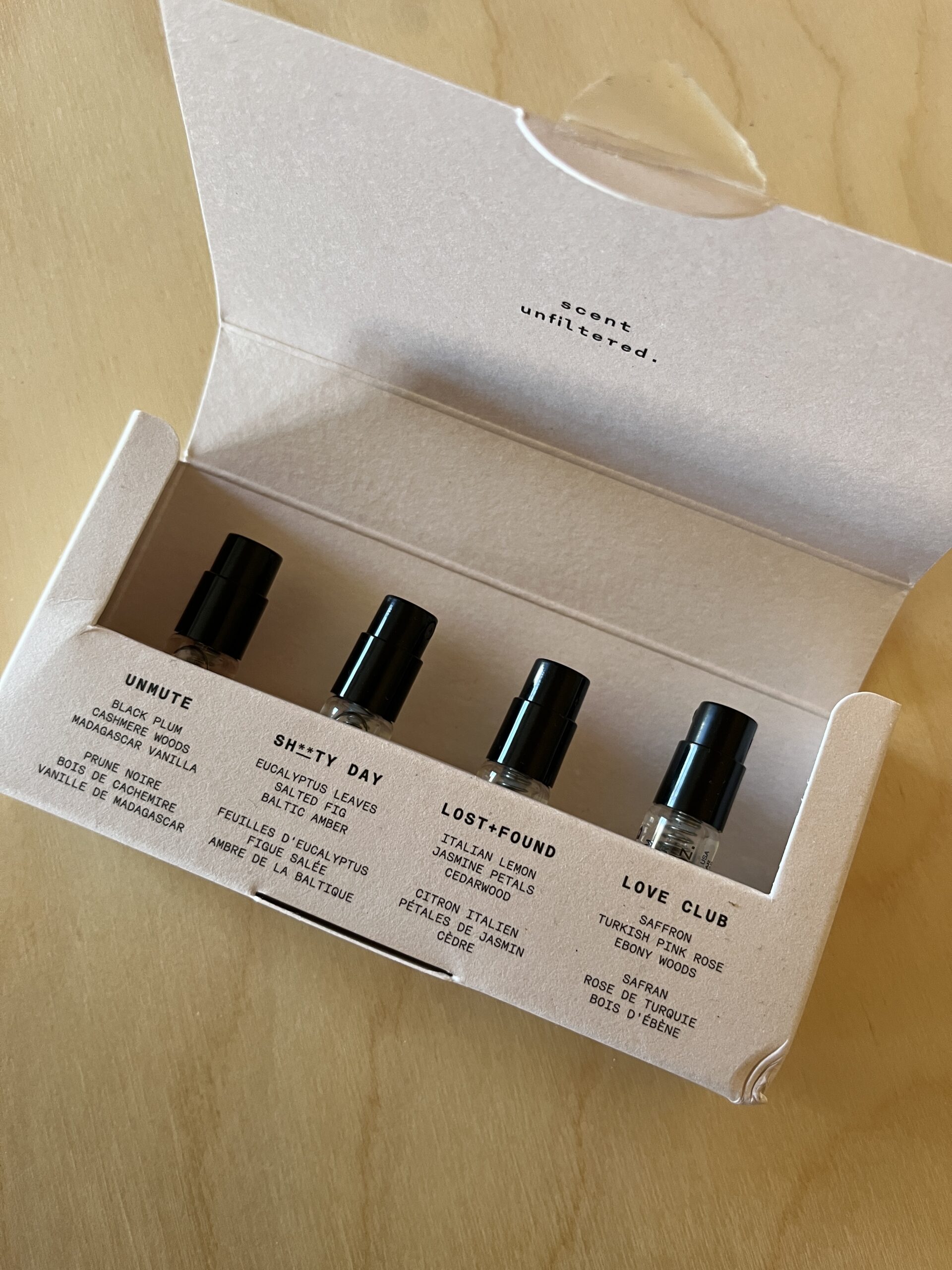 An open cardboard box displaying four small perfume bottles labeled: Unmute, Shitty Day, Lost & Found, and Love Club.