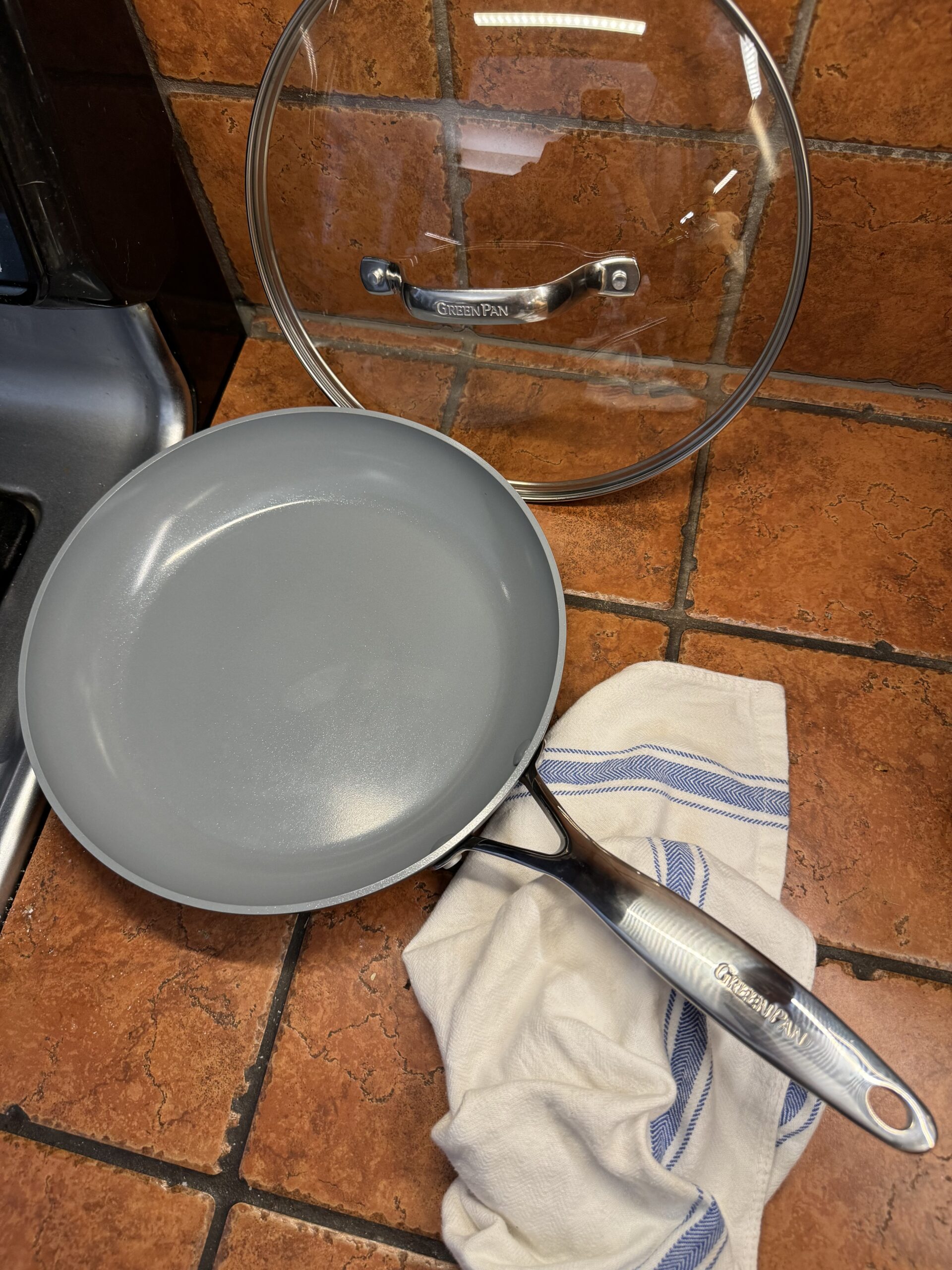 A grey non-stick frying pan with a glass lid, placed on a tiled kitchen countertop next to a white and blue striped dish towel.