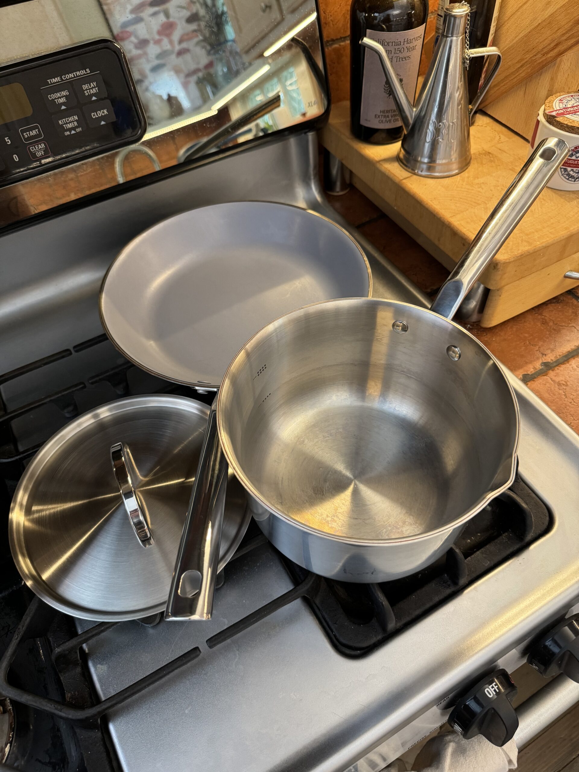 A saucepan with lid and a frying pan, both made of stainless steel, are positioned on a gas stove burner. Nearby are two bottles with metallic pour spouts and some kitchen counter items.