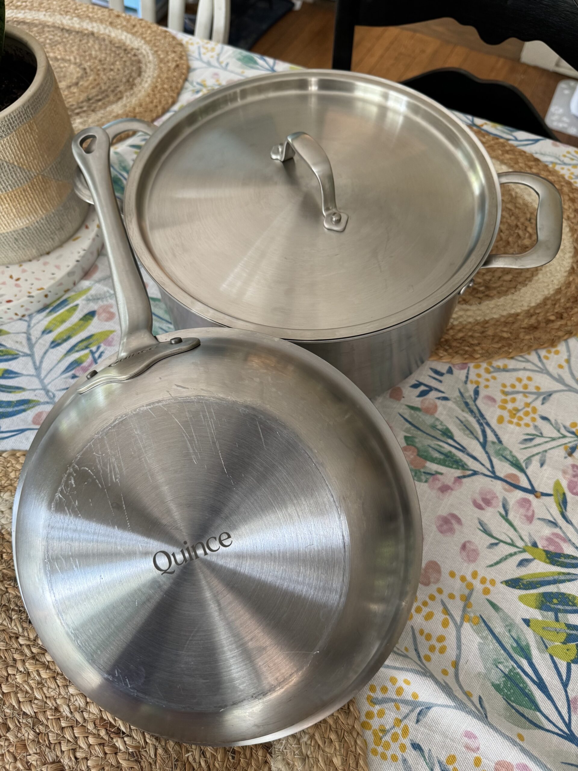 A stainless steel frying pan with "Quince" engraved on its base, placed next to a lidded stainless steel pot on a floral-patterned tablecloth.