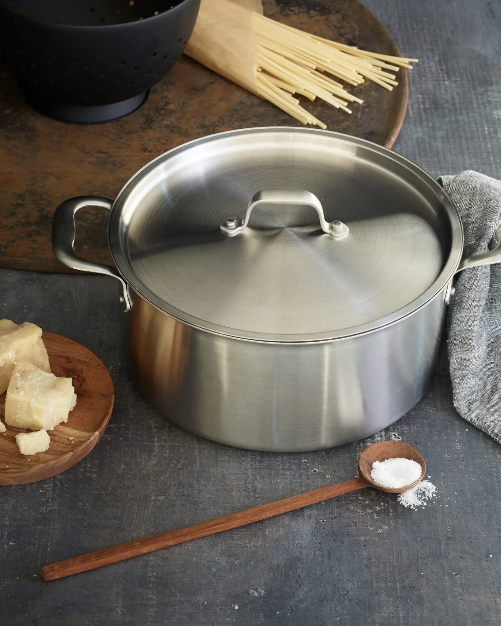 A stainless steel pot with a lid is on a kitchen counter next to a wooden plate with cheese, a small wooden spoon with salt, uncooked spaghetti, and a gray cloth.