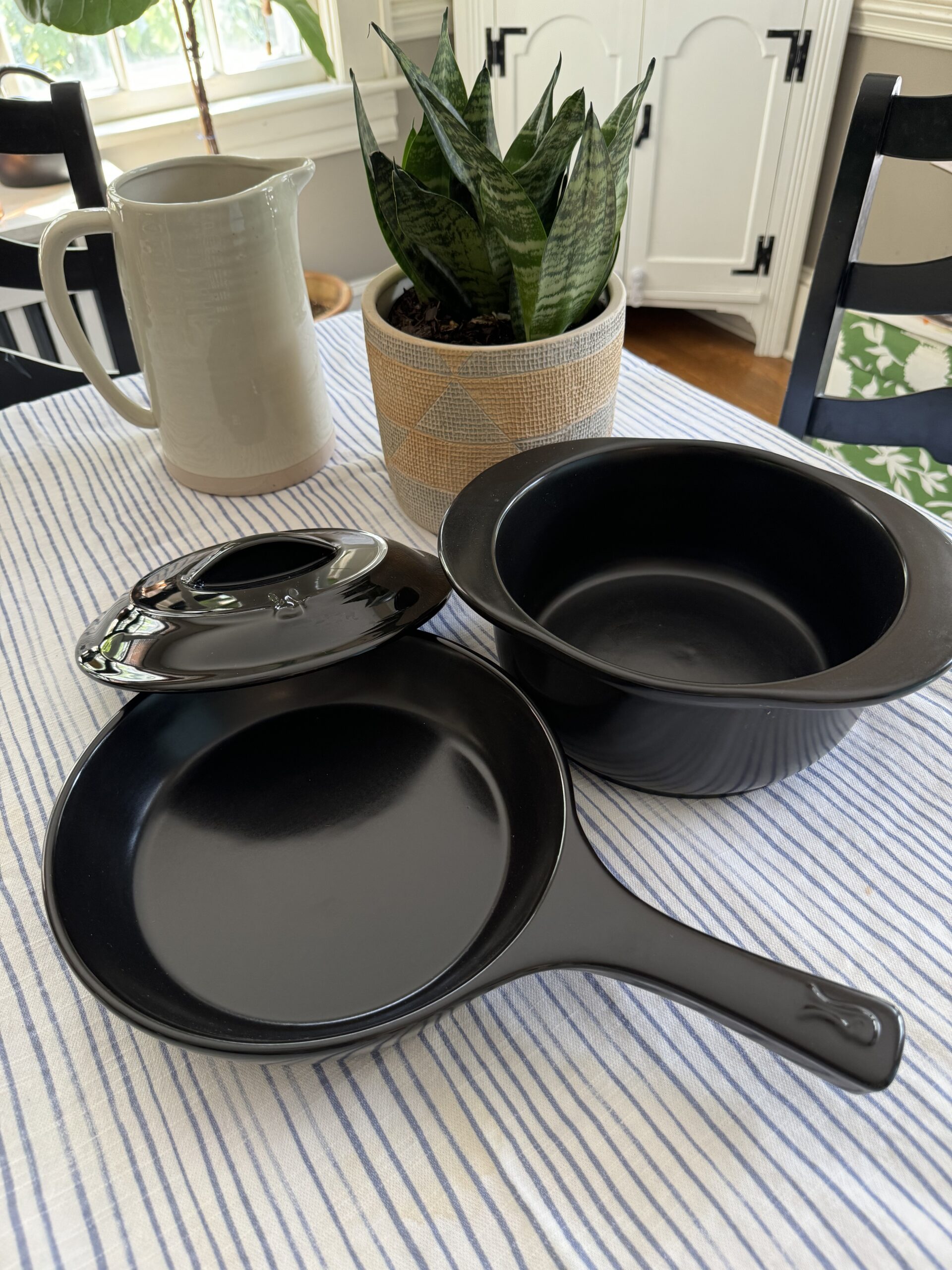 Black frying pan, a pot with a lid, a potted plant, and a ceramic pitcher on a white and blue striped tablecloth.