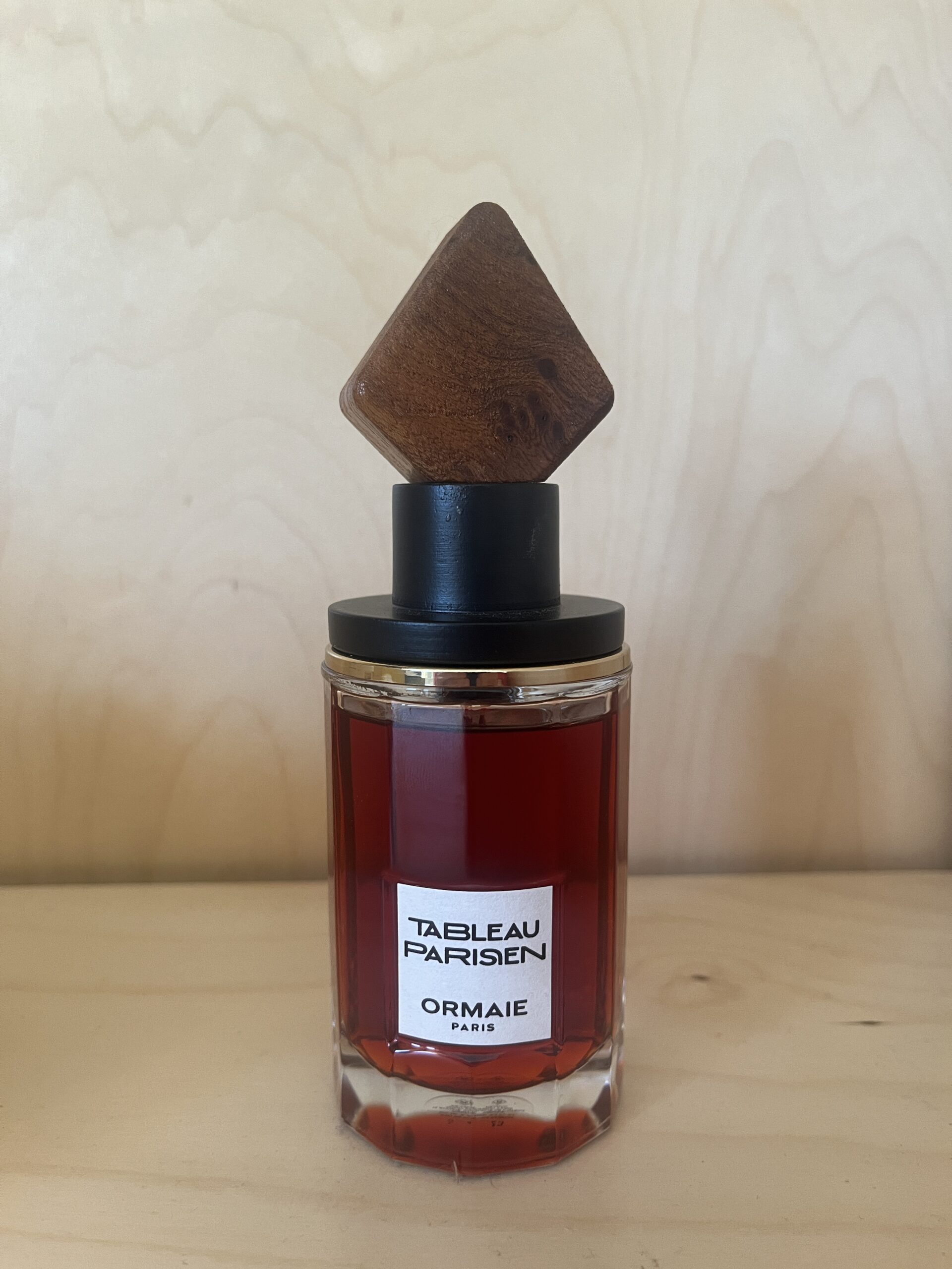 A bottle of ORMAIE Paris perfume with a red liquid, featuring a wooden, square-shaped cap, on a light wood surface.