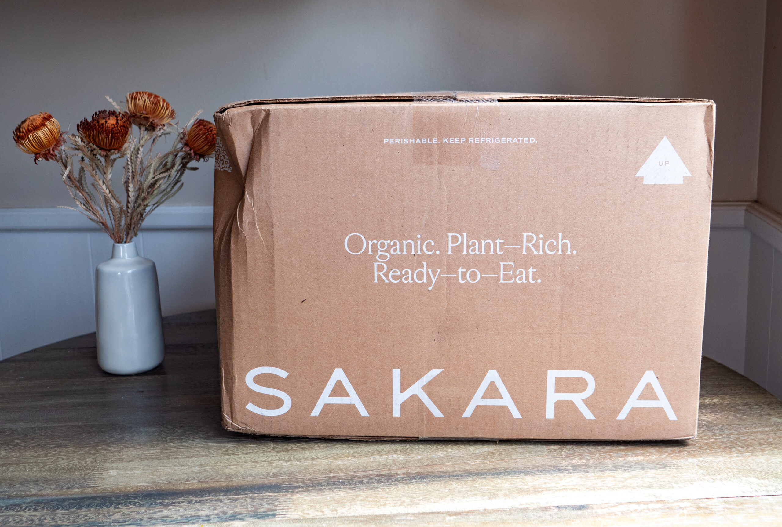 A brown cardboard box labeled "SAKARA" with text "Organic. Plant-Rich. Ready-to-Eat." sits on a wooden table next to a white vase holding dried flowers.