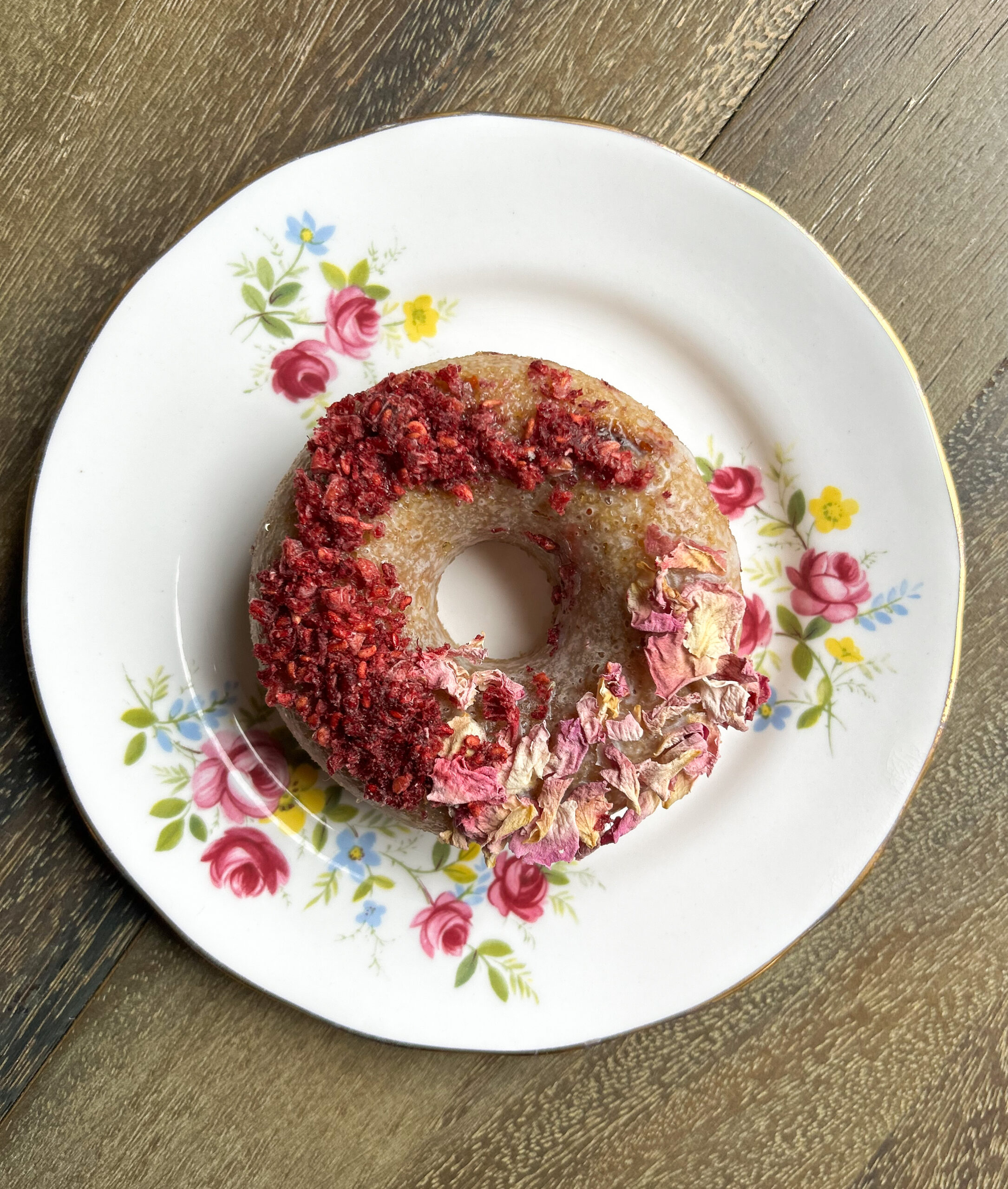 A donut topped with red and pink dried flowers on a floral-patterned plate, set on a wooden surface.