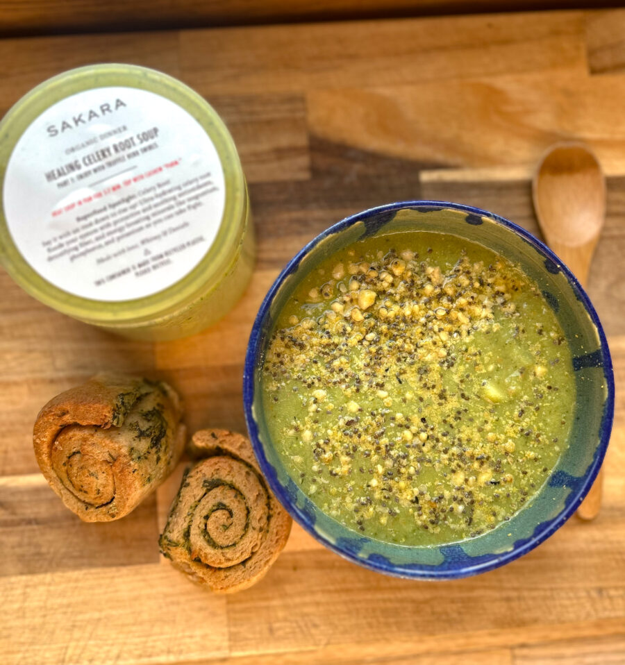 A bowl of green soup with sprinkled seeds on top, two bread rolls, and a container labeled "Healing Green Broth Soup" on a wooden surface with a small wooden spoon.