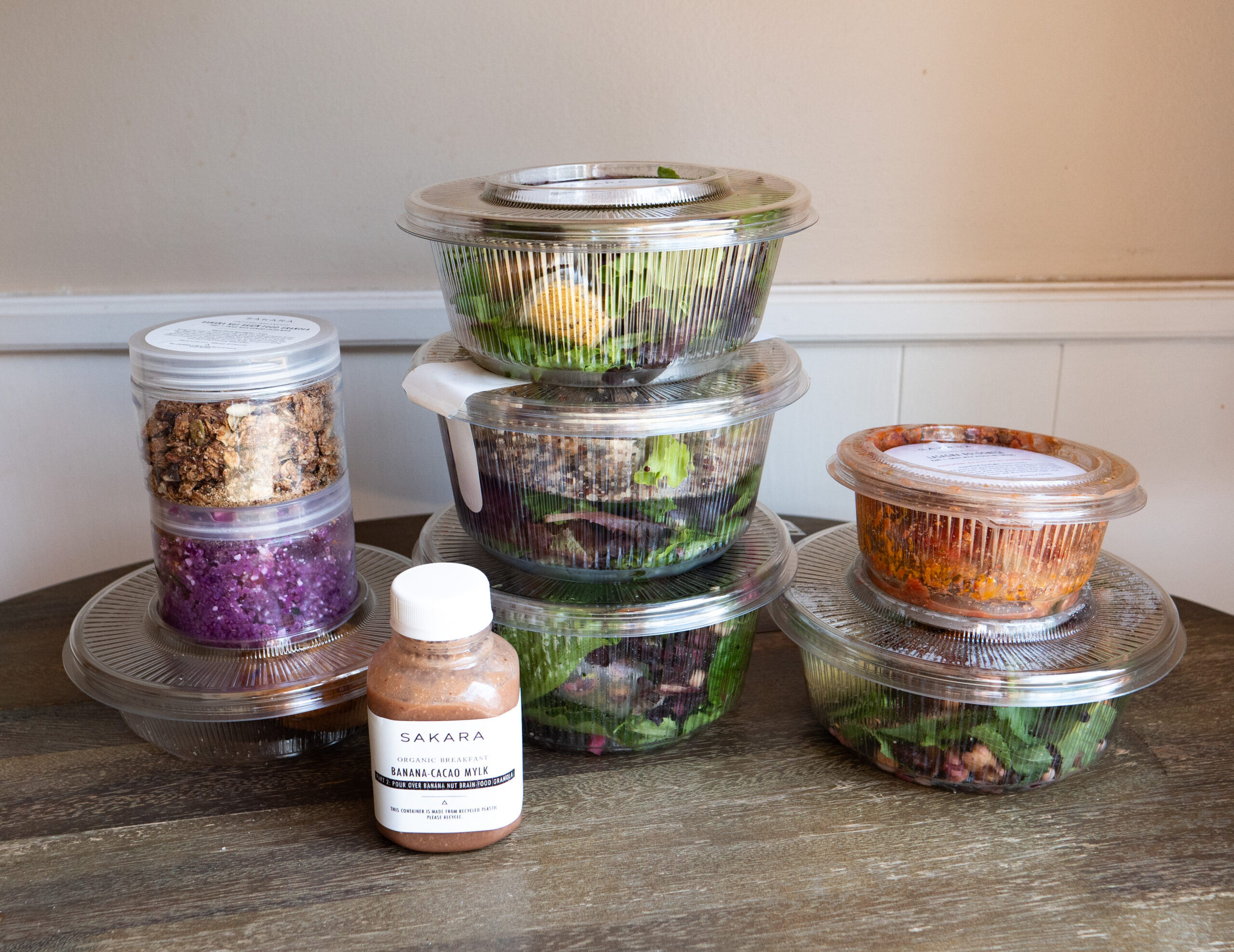 A wooden table with assorted pre-packaged meals in plastic containers and a bottled drink labeled "Sakara." The meals include salads, granola, and other food items.