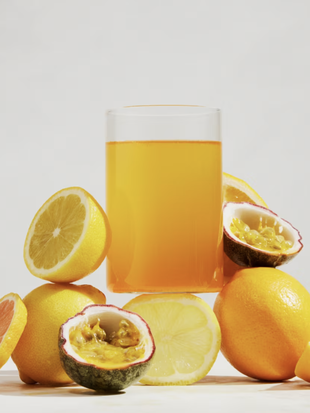 A glass of orange juice surrounded by various citrus fruits, including lemons, oranges, and halved passion fruits, on a white background.