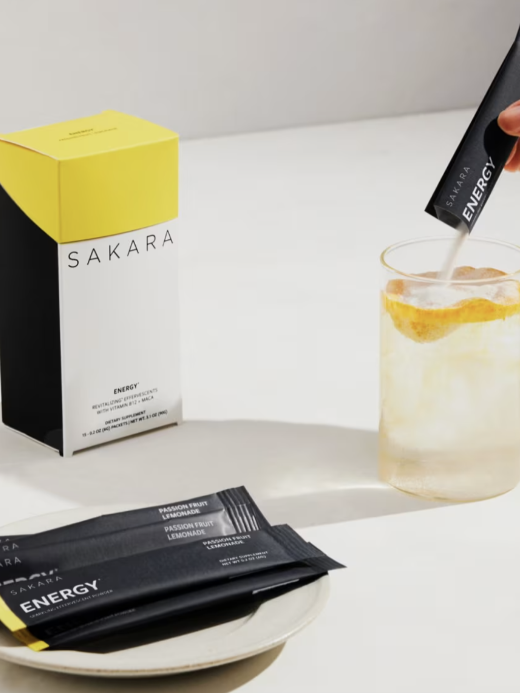 A hand pours a packet of Sakara Energy powder into a glass of lemon water. A box and three more packets are on a white surface.