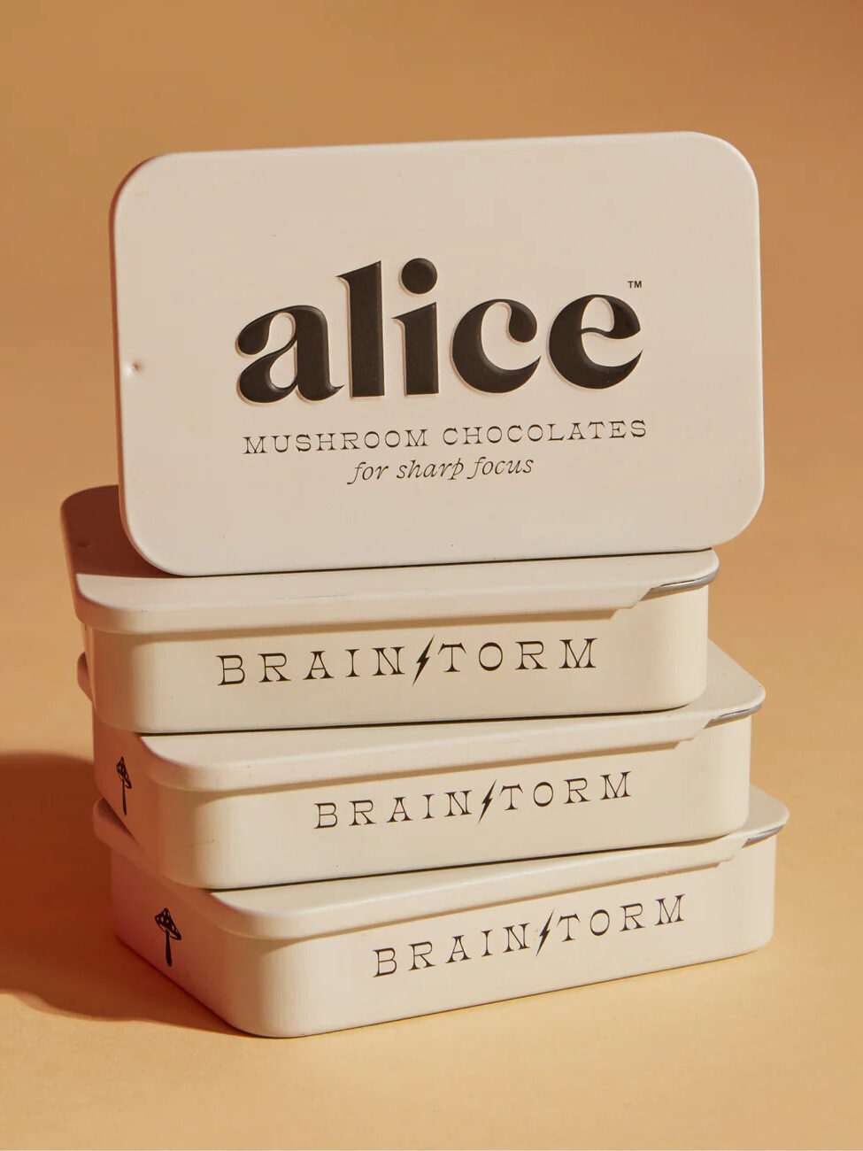 A stack of four white tins labeled "alice MUSHROOM CHOCOLATES for sharp focus" and "BRAIN STORM" on a beige background.