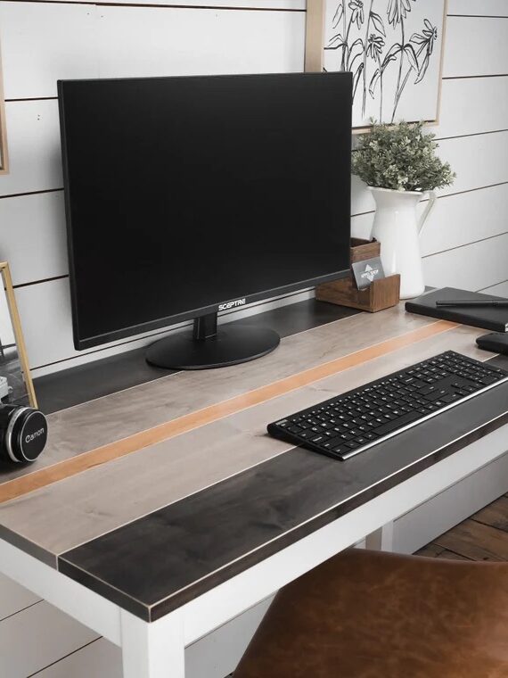 A tidy workspace featuring a monitor, keyboard, camera, and a potted plant on a wooden desk with a chair. Framed pictures and decorative items are on the wall and tabletop.